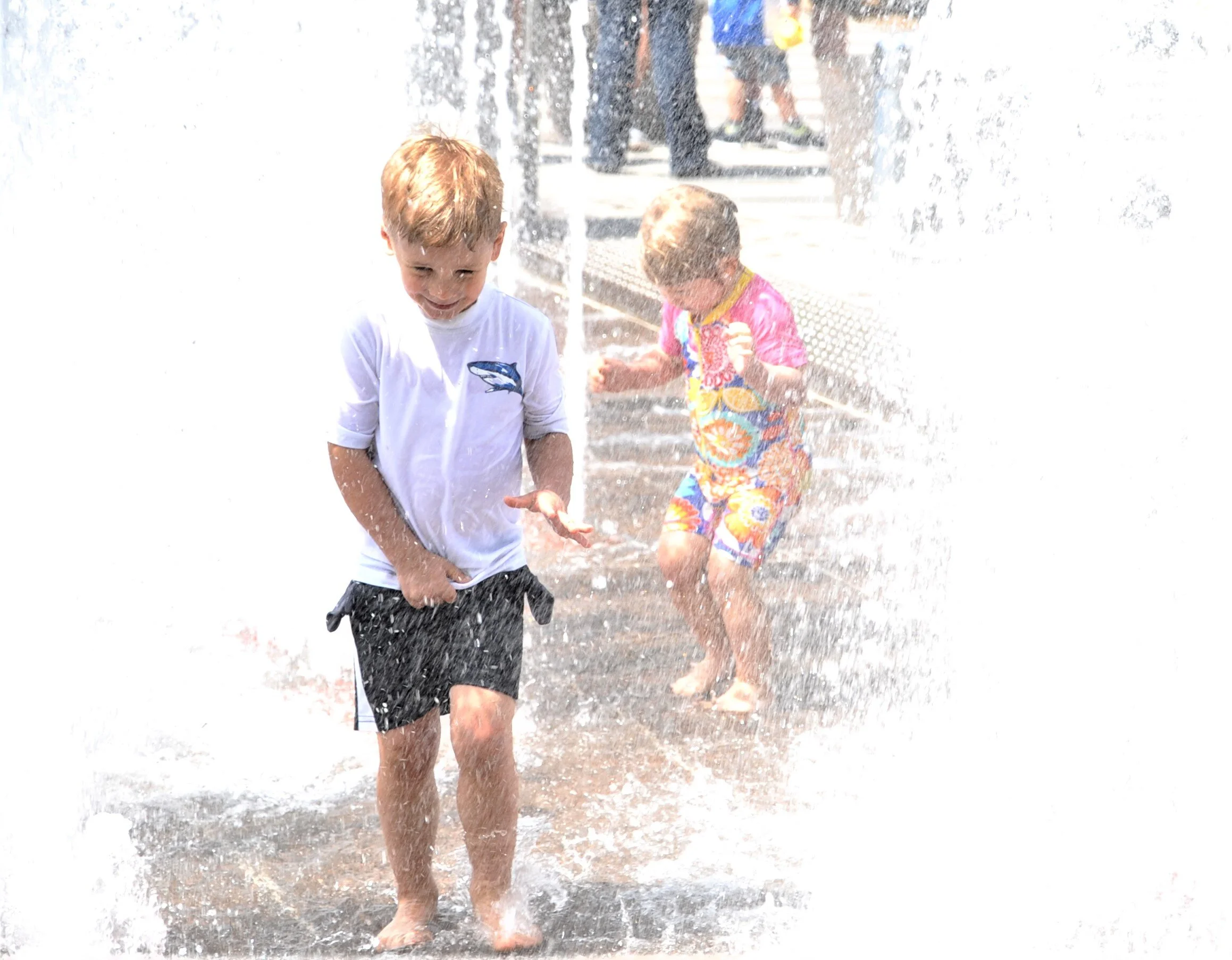 Two children playing in a fountain, one boy smiling and walking through water, and a girl behind him in colorful clothing, both splashing in water on a sunny day.
