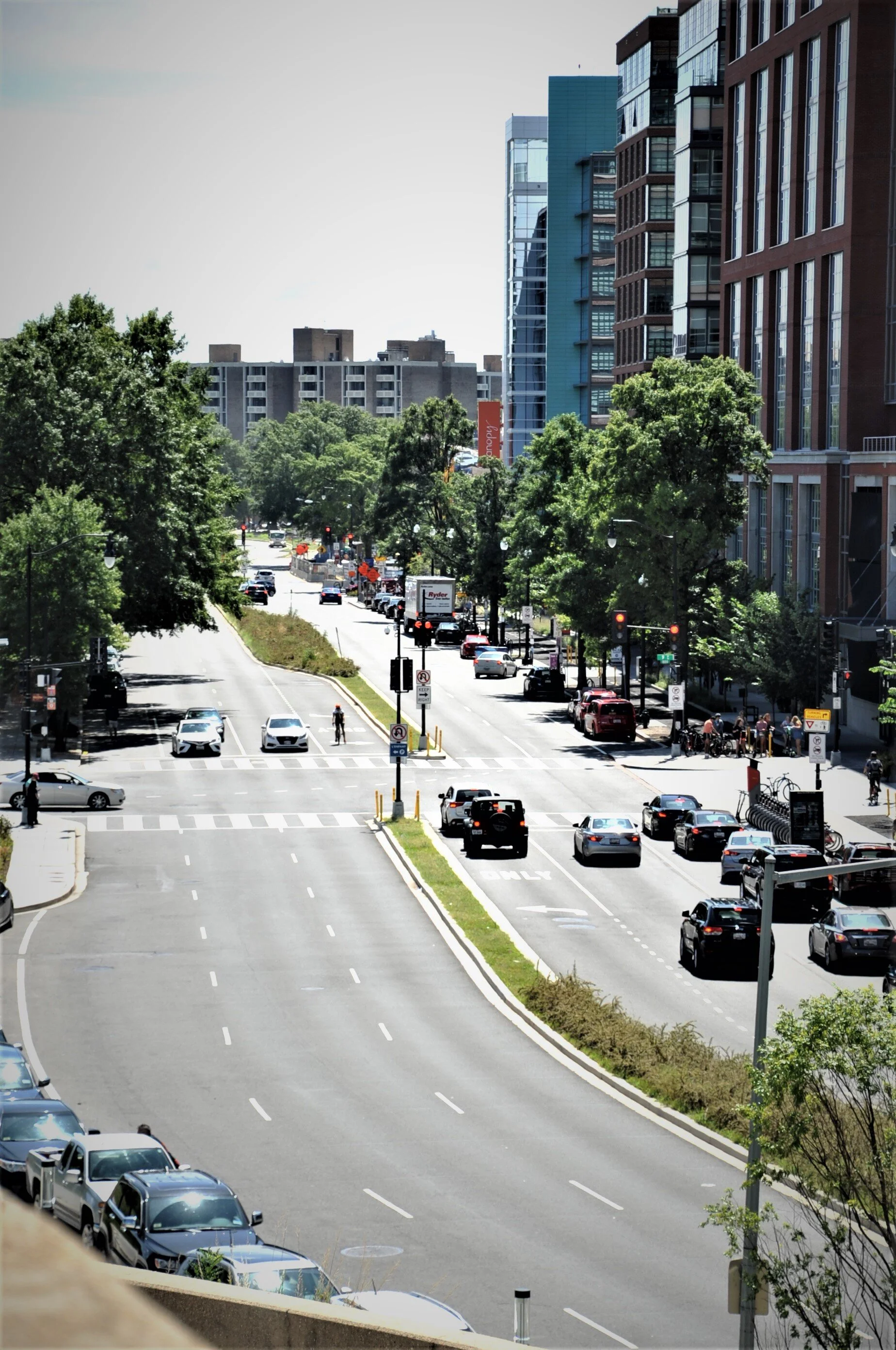City street with cars, pedestrians, trees, and tall modern buildings under a clear sky.