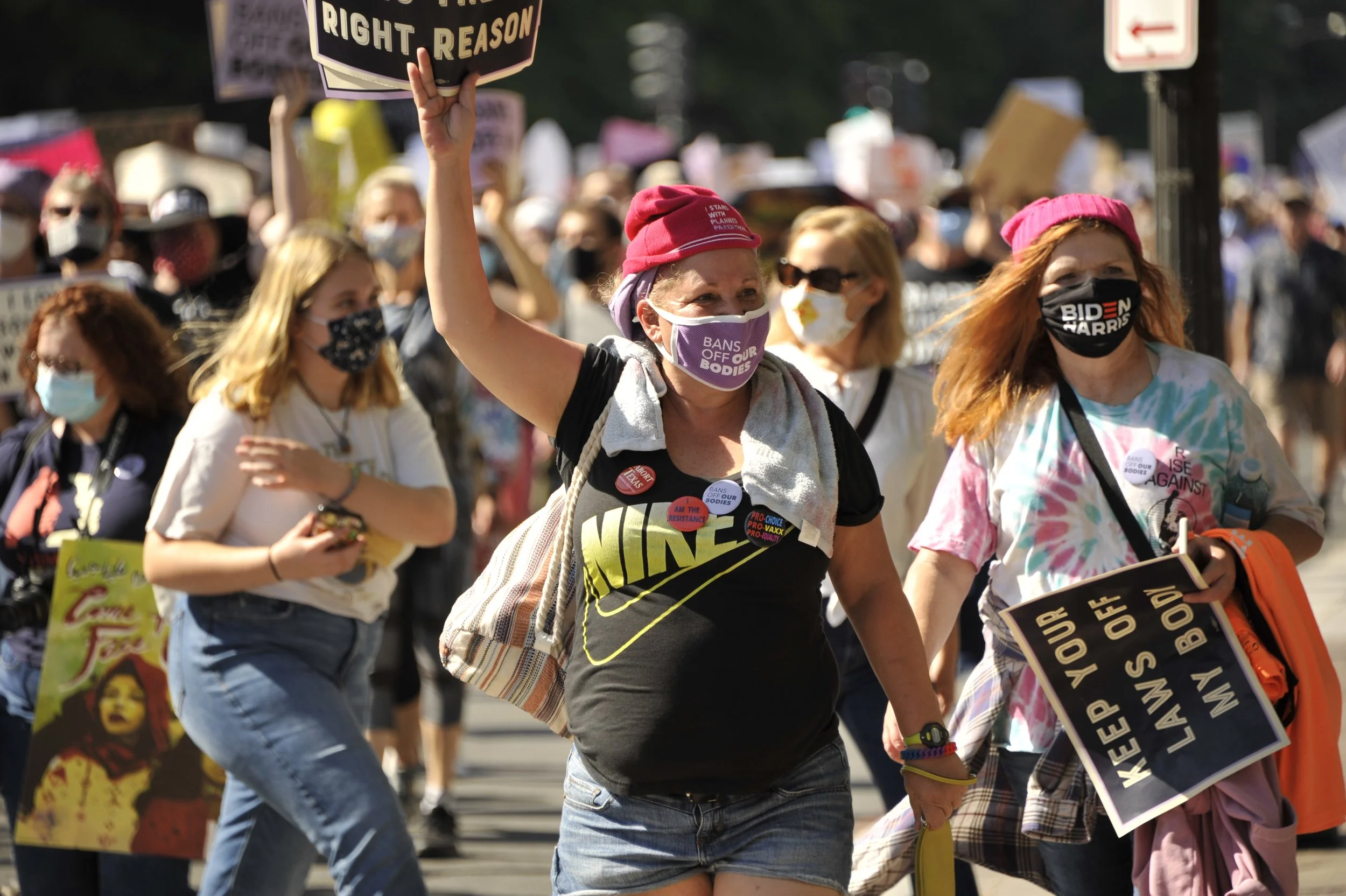A woman at a protest wearing a purple face mask with the words "BANS OFF OUR BODIES," holding a sign and surrounded by other protesters, many wearing masks and holding signs.