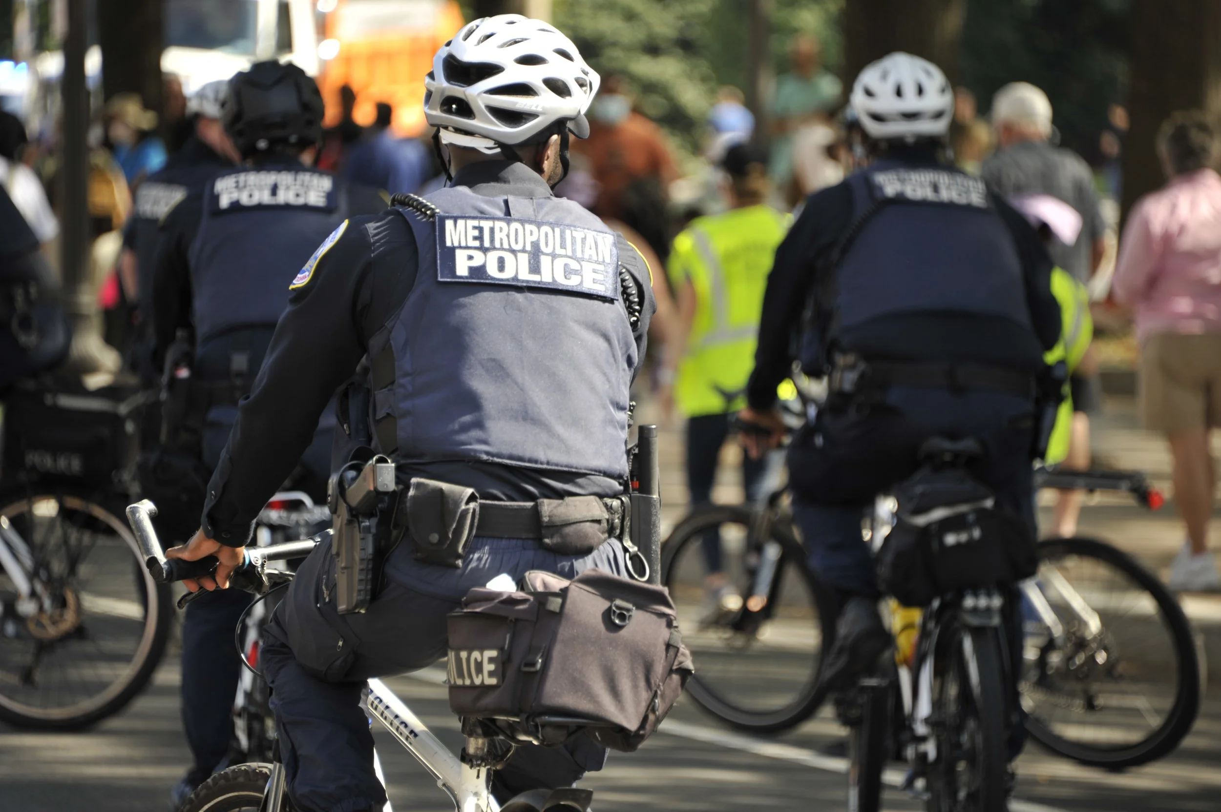 Metropolitan police officers riding bicycles during a public event, with people walking and standing in the background.