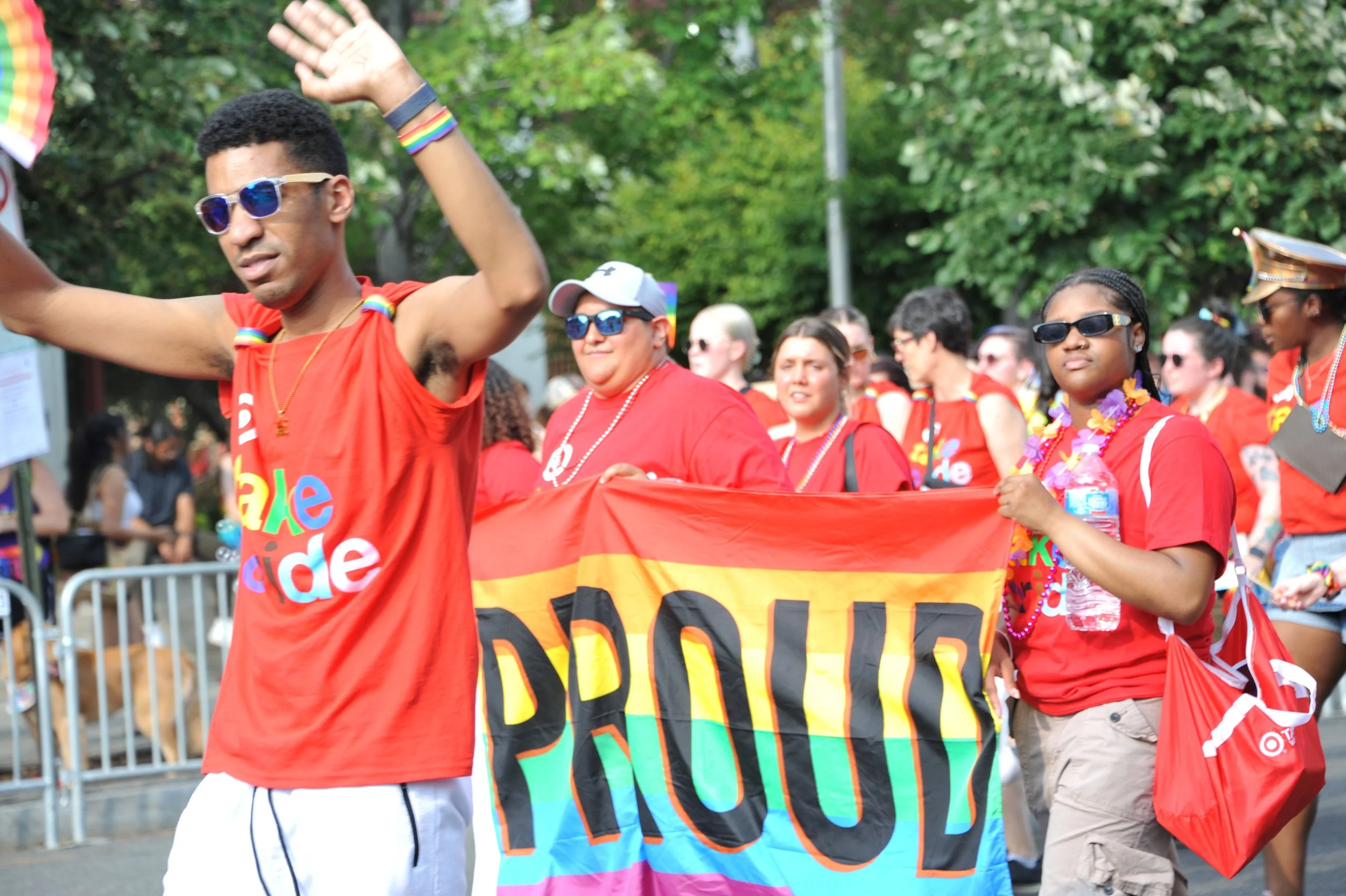 People participating in a pride parade, wearing red shirts with rainbow-colored text, carrying a rainbow flag, and some with rainbow accessories and drinks, walking outdoors with trees in the background.