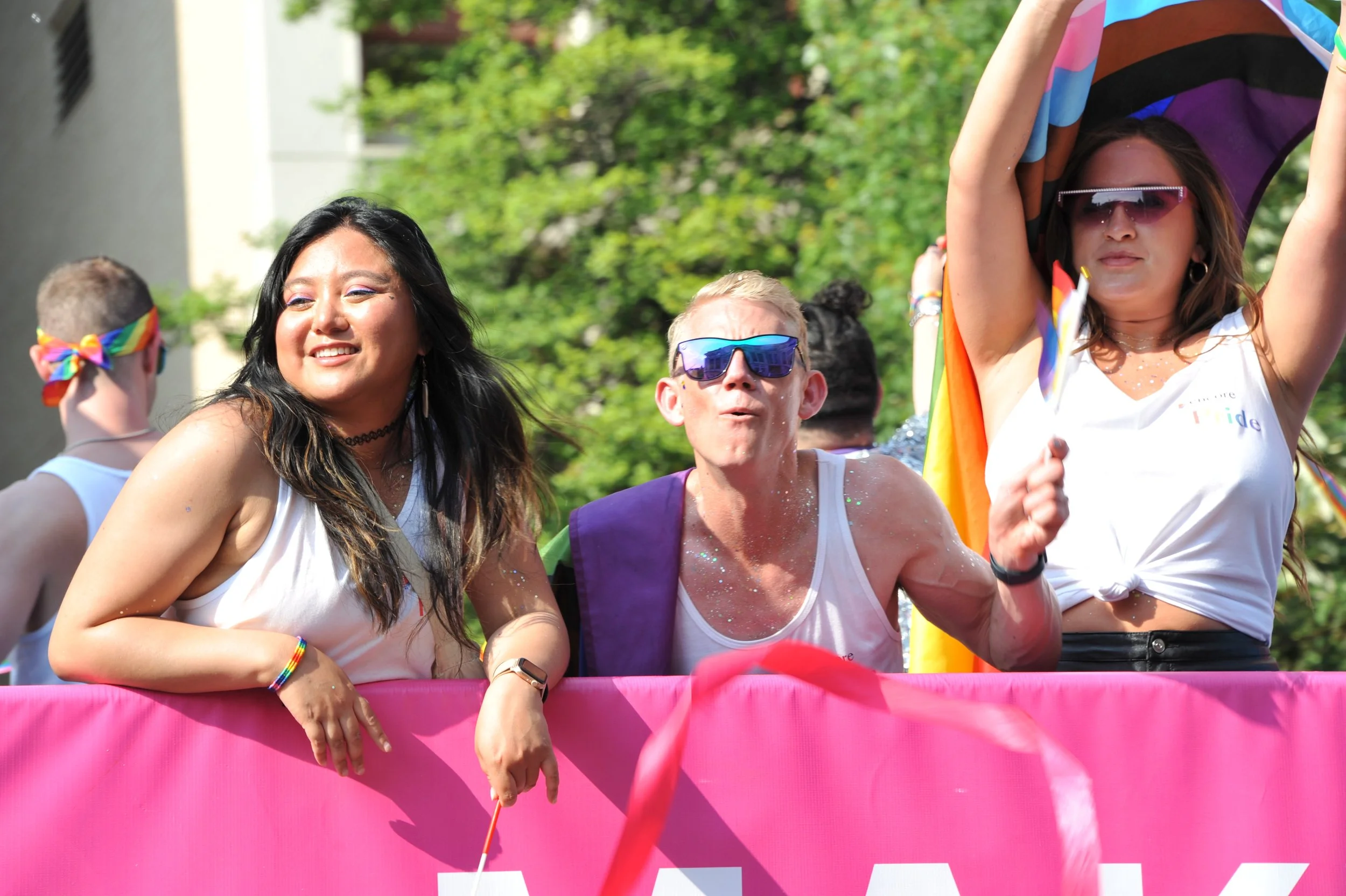 People celebrating at a pride parade, wearing white shirts, some with rainbow accessories, behind a pink barrier, with colorful flags and greenery in the background.
