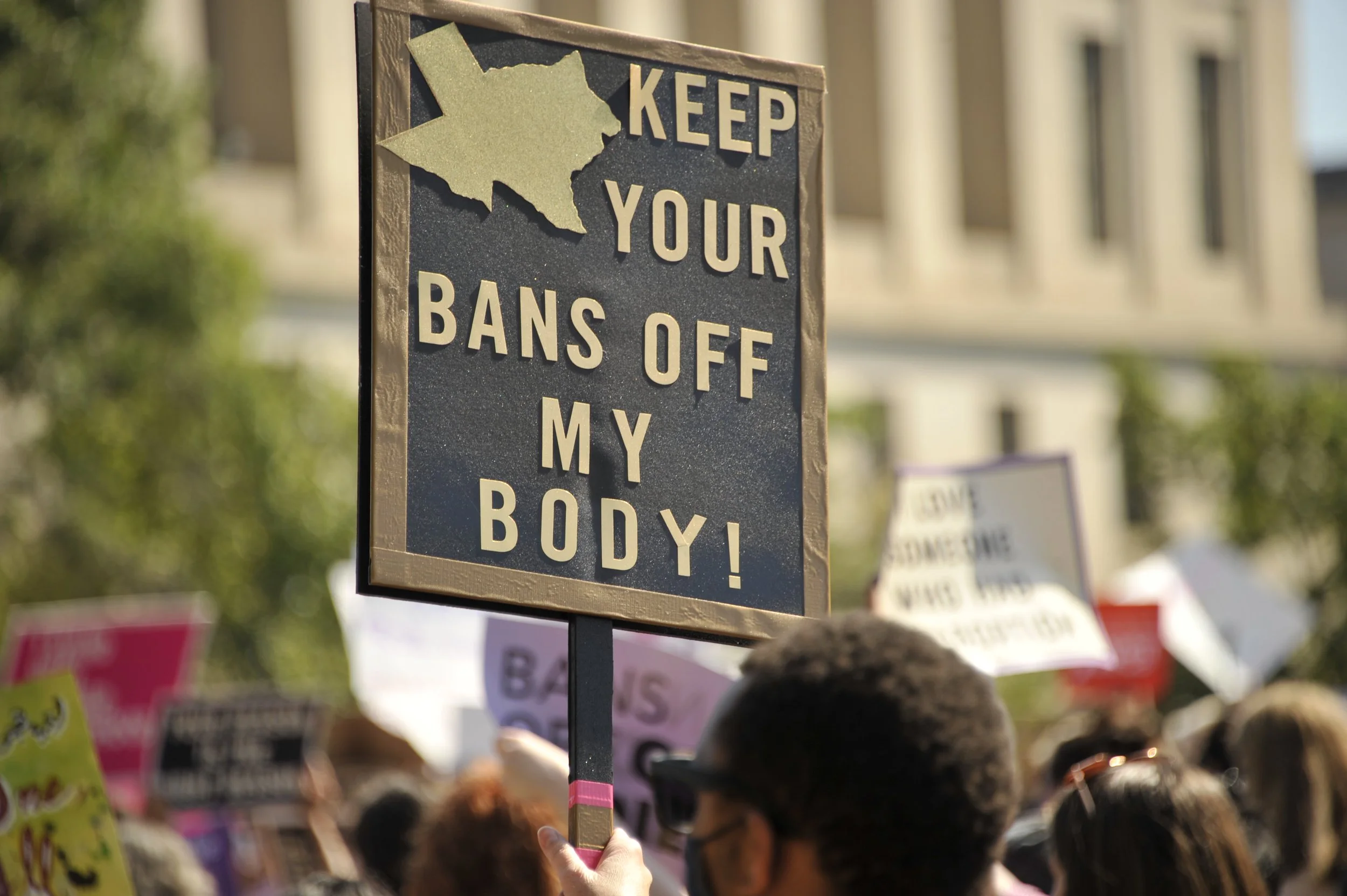 A protest sign reading 'Keep Your Bans Off My Body!' held by a person among a crowd of protesters.