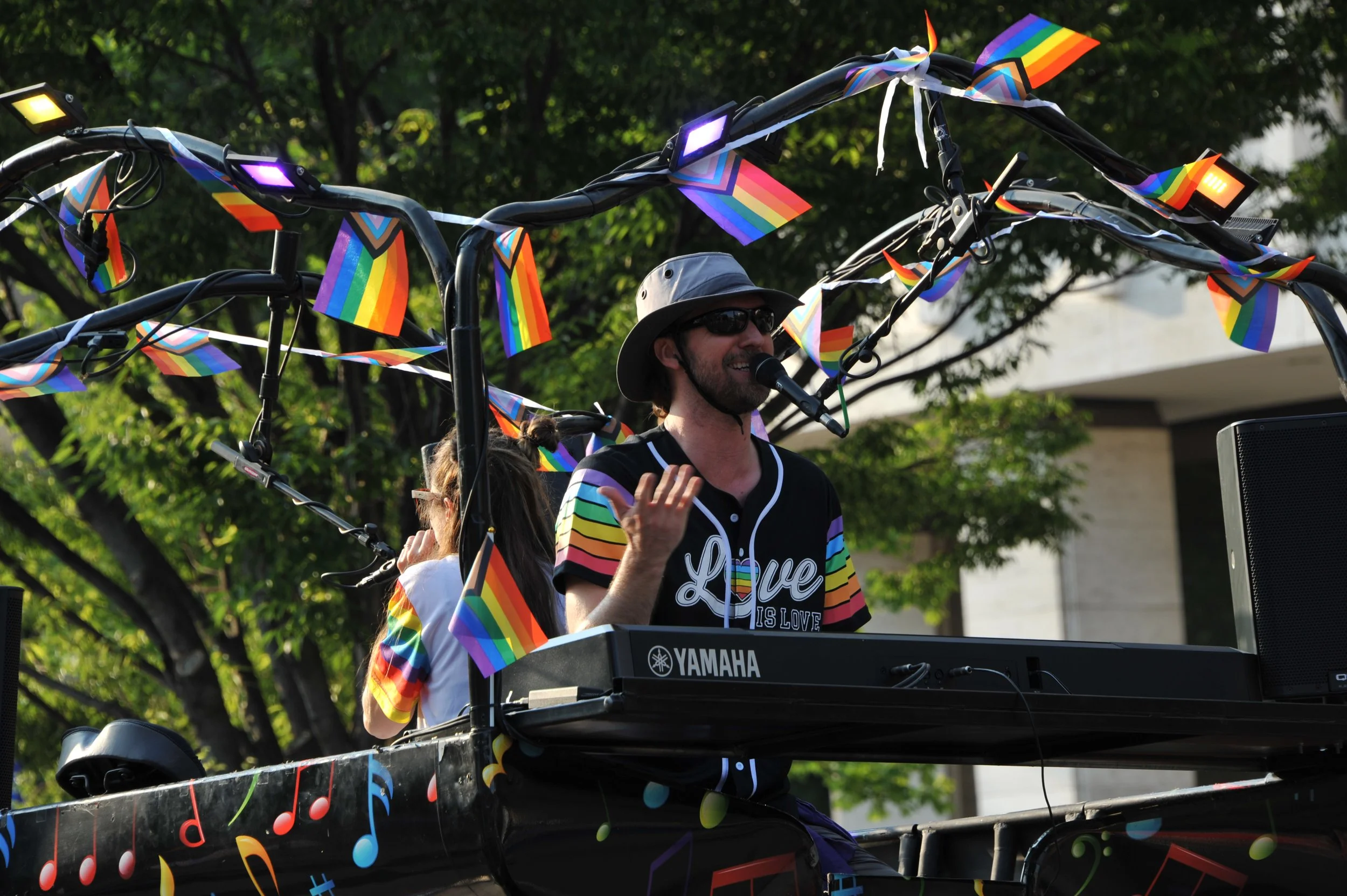 A DJ wearing sunglasses, a hat, and a colorful rainbow-themed shirt, performing at a pride parade or festival on a Yamaha keyboard, decorated with rainbow flags and musical note stickers, outdoors with trees in the background.