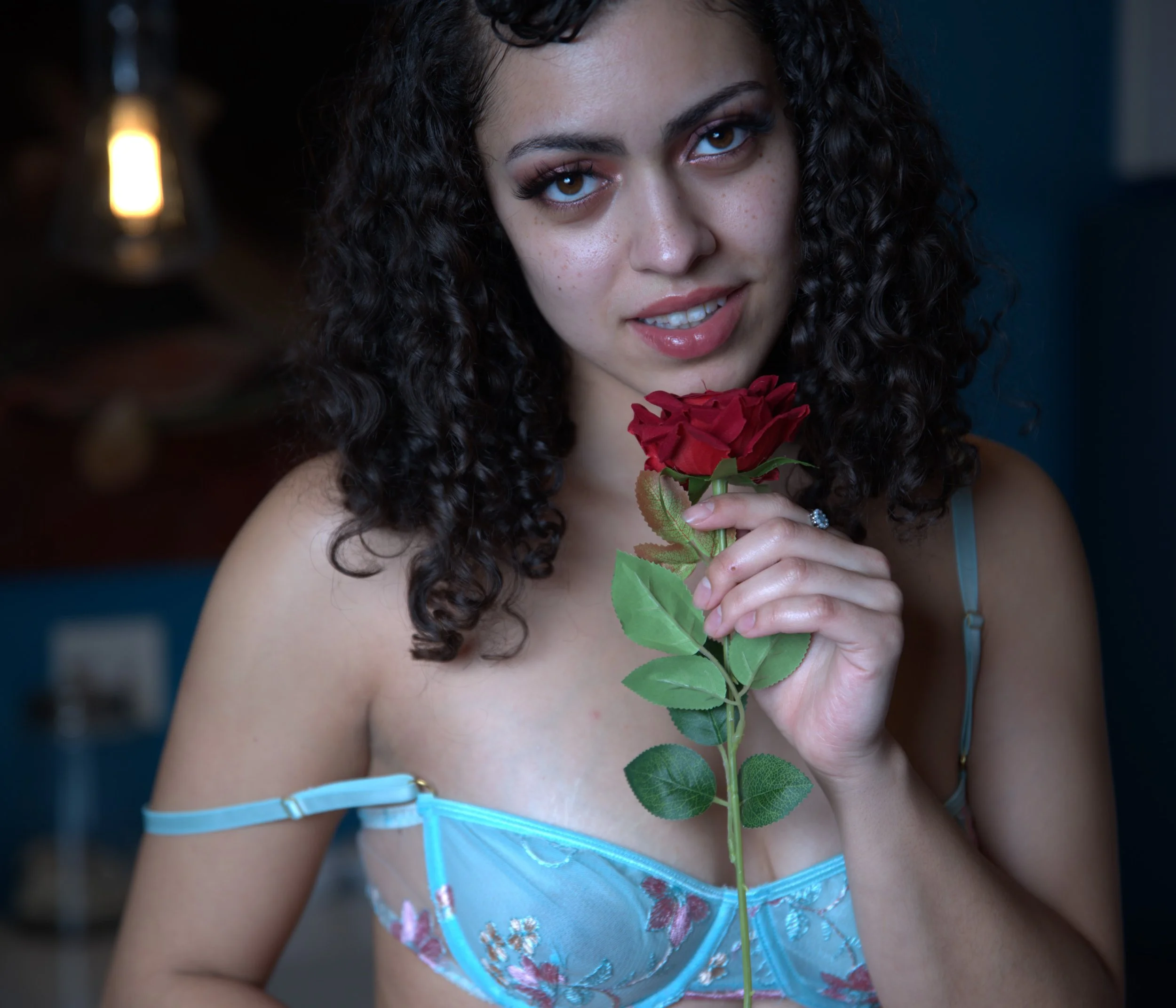 A woman with curly dark hair holding a red rose in front of her chest, smiling softly, wearing a blue floral lingerie top, in a dimly lit room.