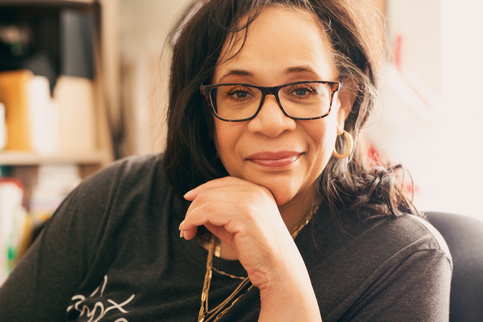 A woman with dark hair, glasses, and hoop earrings is resting her chin on her hand, smiling softly. She is indoors with a blurred background. The lighting suggests natural sunlight.