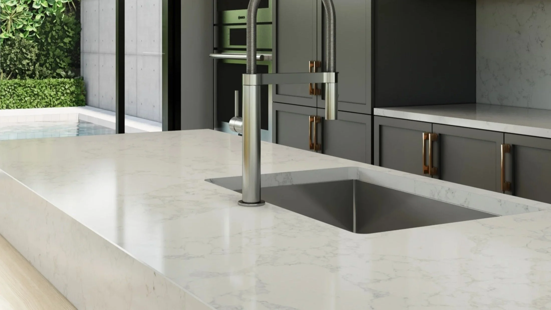 A modern kitchen with a white marble countertop, under-mount sink, and a sleek metal faucet. Gray cabinets with gold handles are visible, with an indoor view opening to an outdoor area with greenery and a pool.