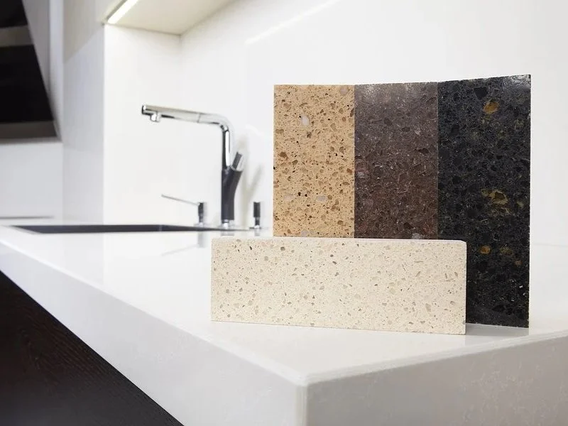 Sample display of four different countertops or stone slabs in beige, brown, dark brown, and black colors against a white countertop and modern kitchen sink.