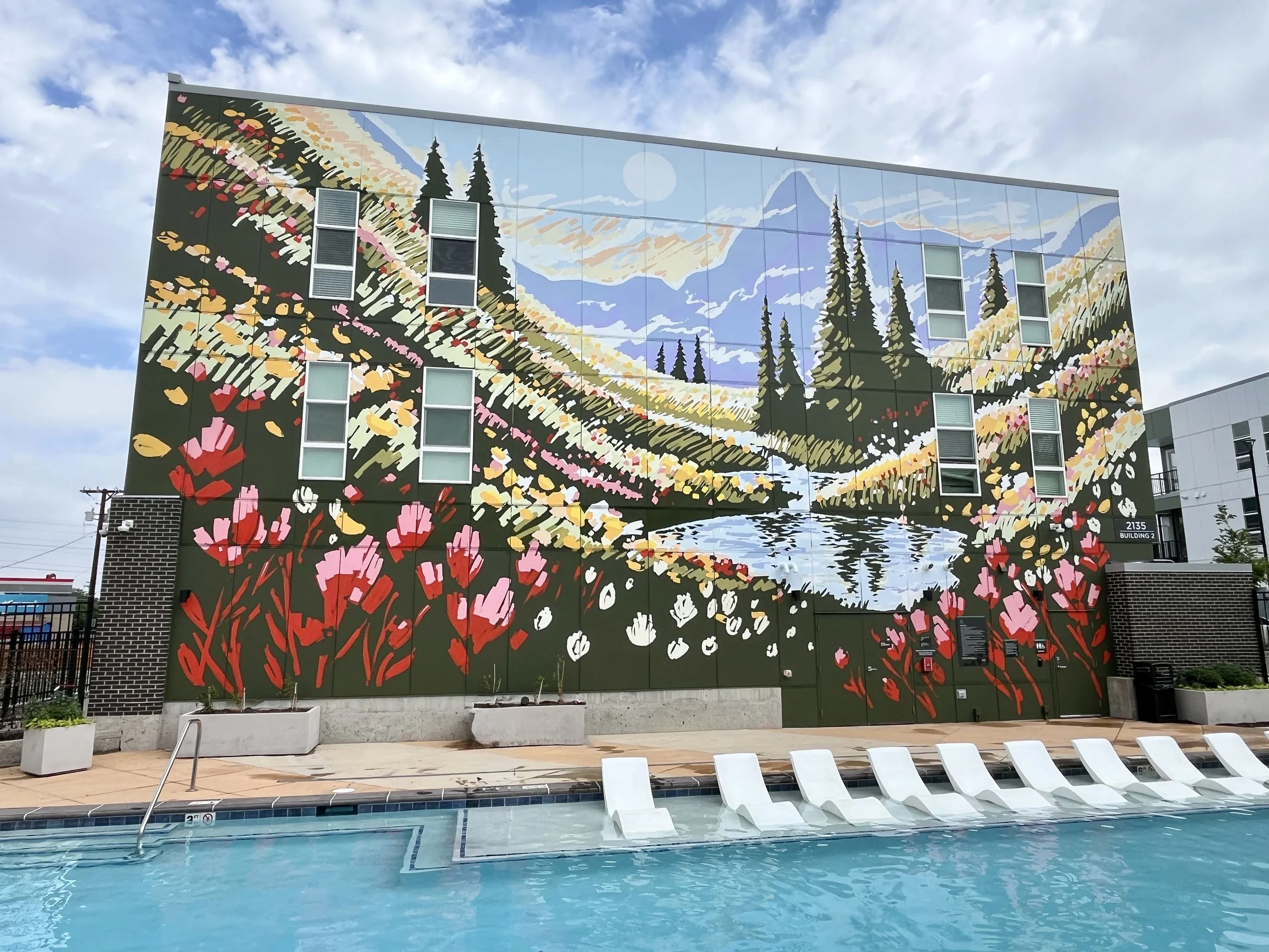 A colorful mural of a mountainous landscape with trees, hills, and flowers is painted on the side of a building above a swimming pool.