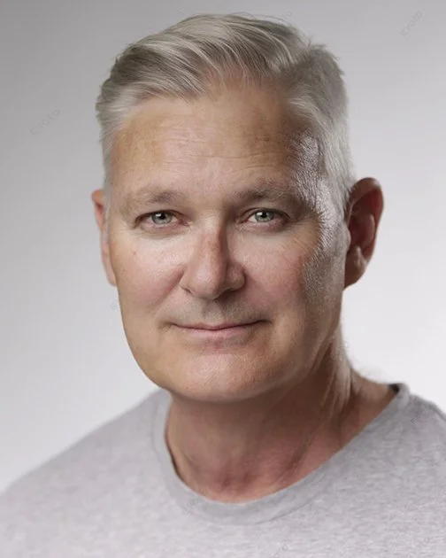 A middle-aged man with gray hair and blue eyes smiling at the camera, wearing a light gray T-shirt against a plain light gray background.