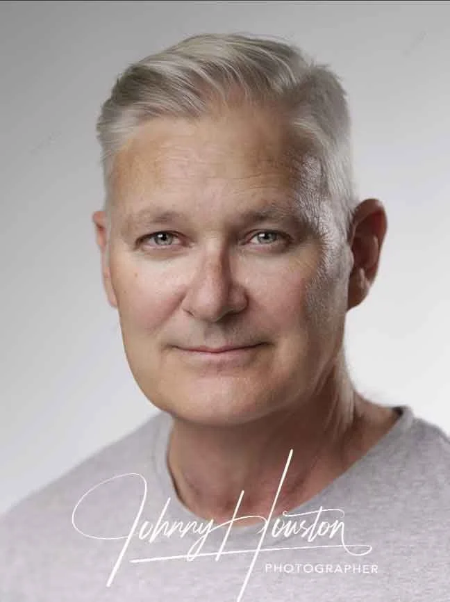 Headshot of a middle-aged man with short blonde hair, fair skin, and light-colored eyes, smiling slightly, wearing a gray shirt, with the photographer's signature 'Johnny Houston' and 'Photographer' written at the bottom.