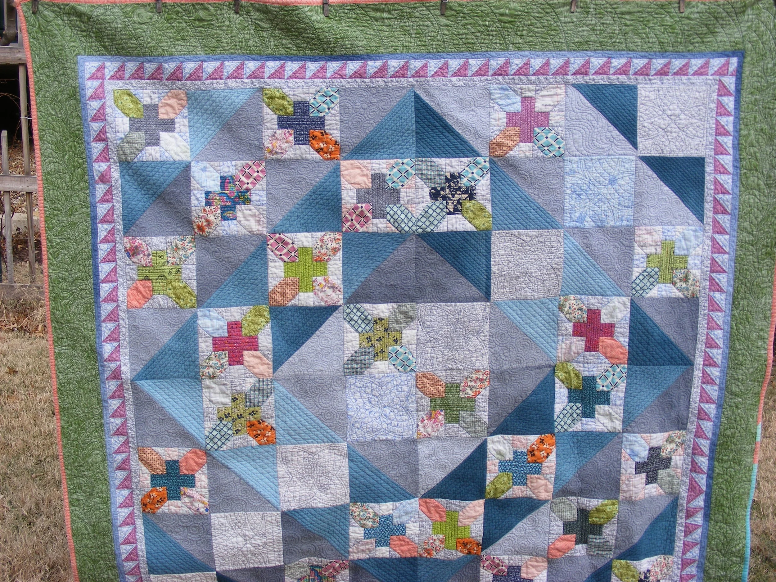 A colorful patchwork quilt with various geometric patterns and cross designs, featuring shades of blue, green, purple, and pink, bordered with a green edge.