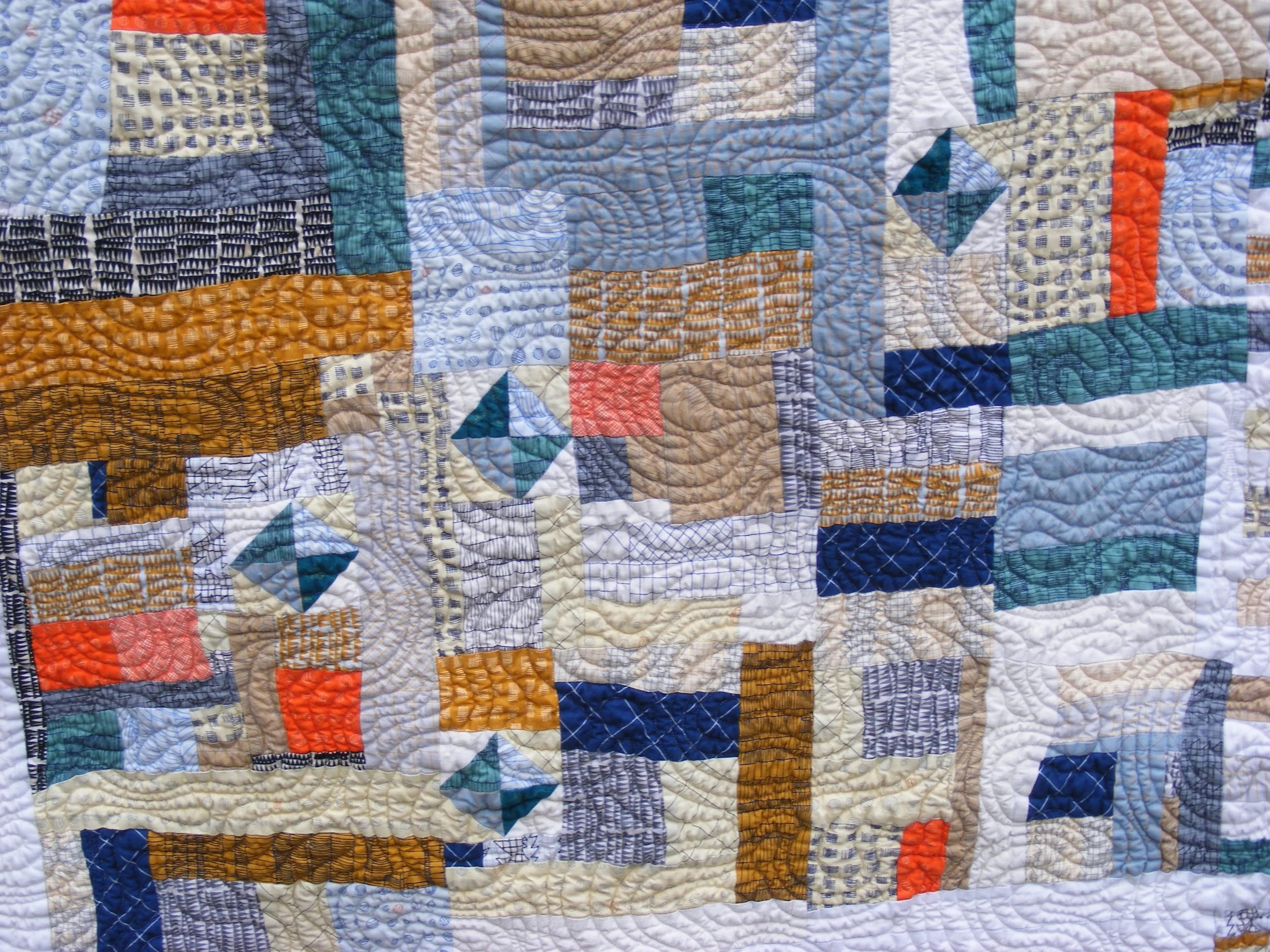 Abstract quilt with multicolored geometric patterns, including rectangles and squares, in shades of blue, orange, brown, and cream. Features intricate stitching and varied textures.