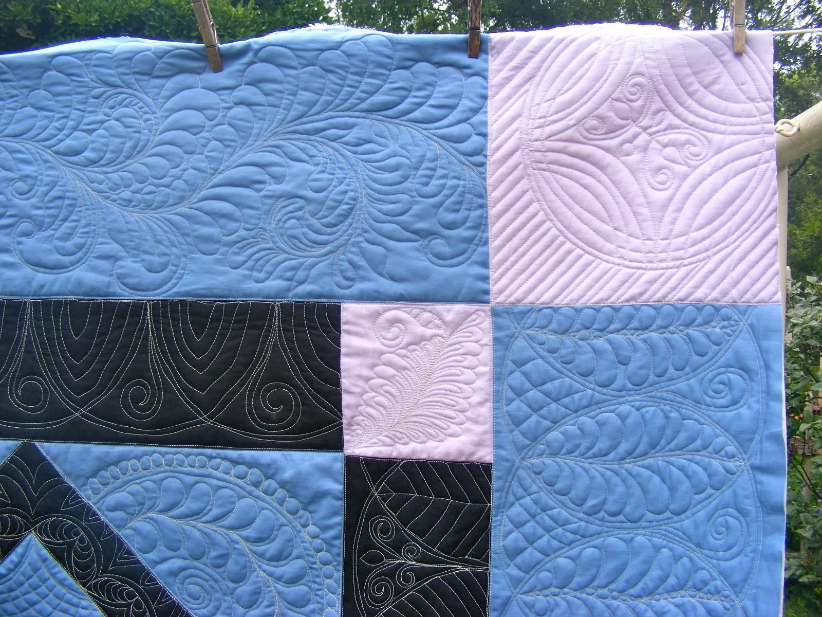 Patchwork quilt with intricate quilting patterns, featuring blue, black, and pale pink sections, hanging outdoors.