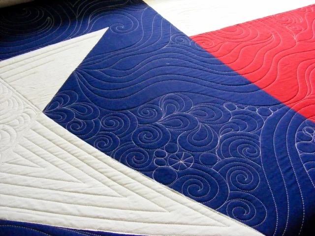 Close-up of a quilt featuring a large white star on a blue background, with intricate swirling and geometric stitch patterns. The design also includes a section of red fabric with similar stitching.