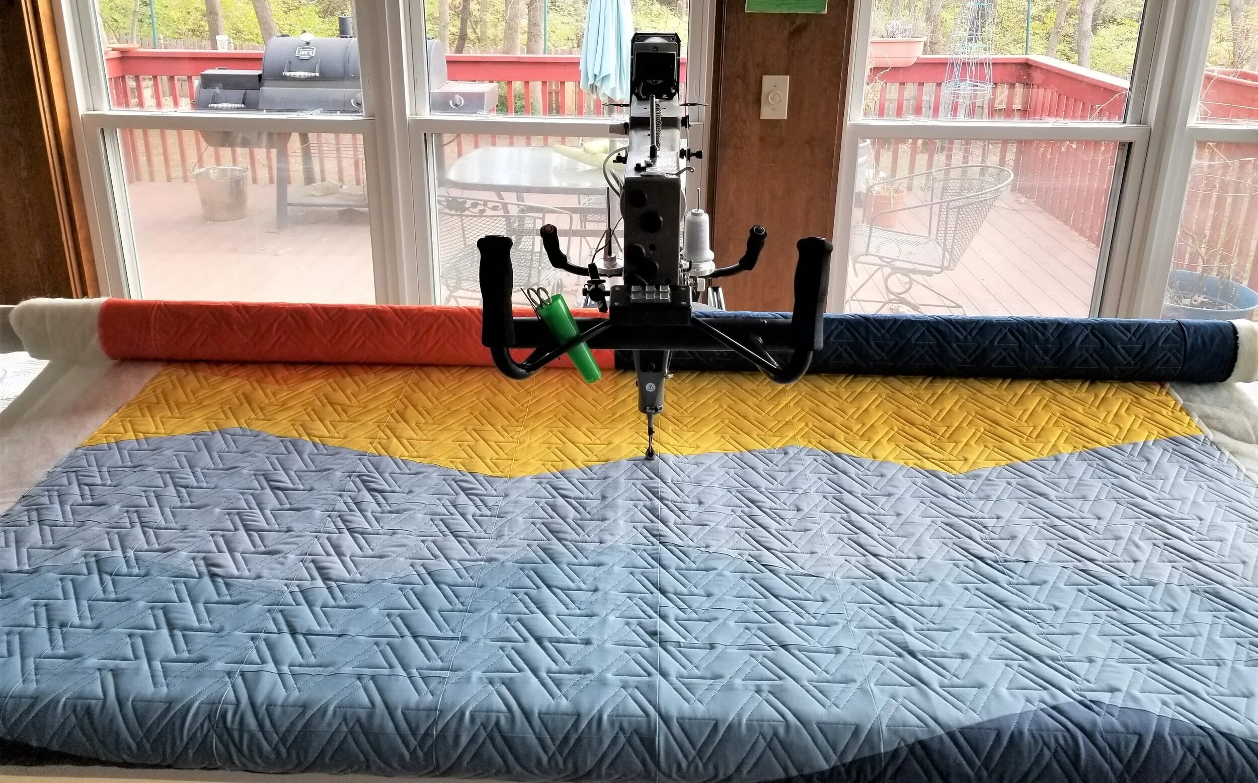 Longarm quilting machine with multicolored quilt in progress, blue, yellow, and orange sections visible. Outdoor patio view in background.