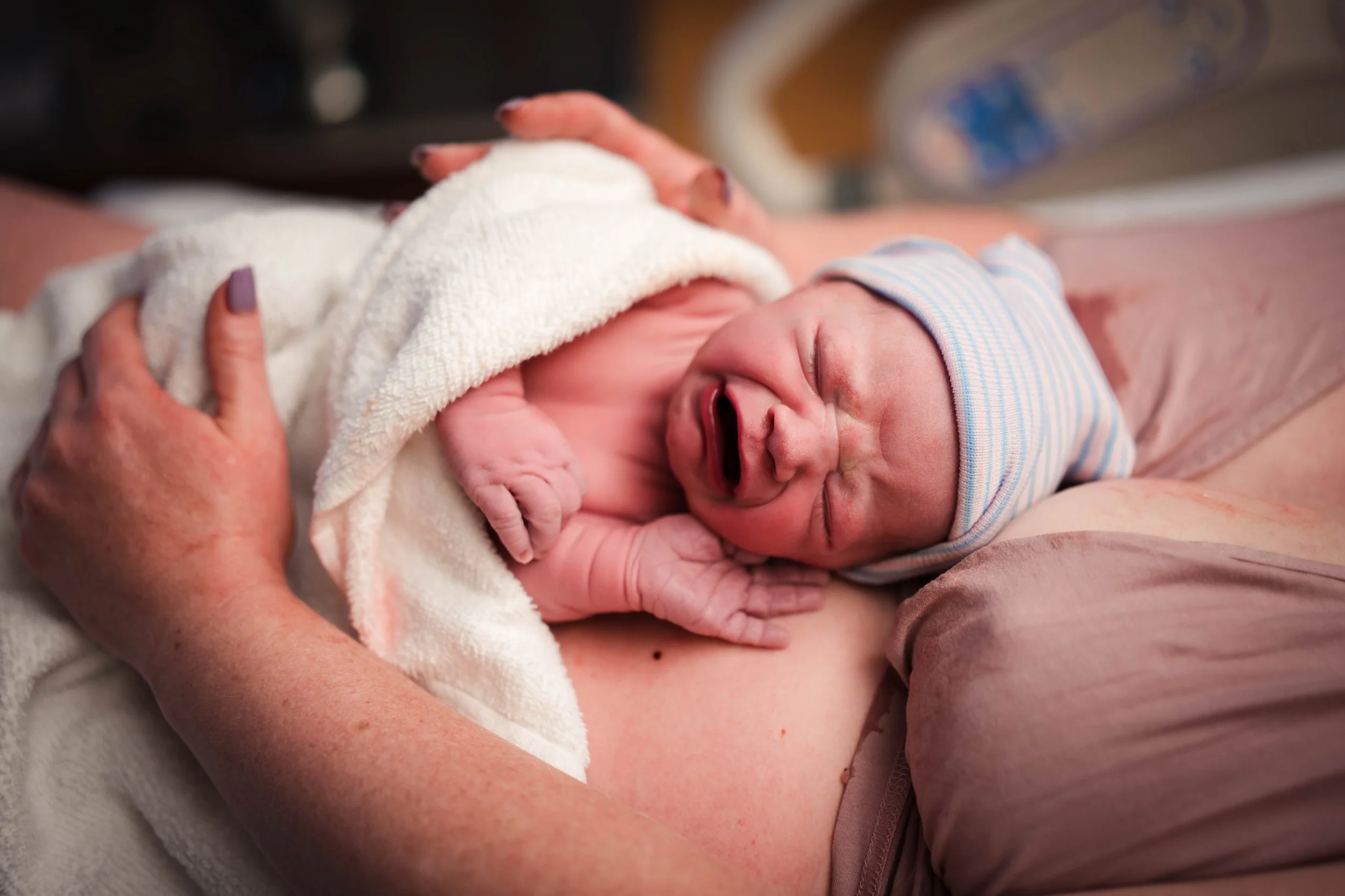 A newborn baby wrapped in a blanket and wearing a striped hat, crying while being held by an adult.