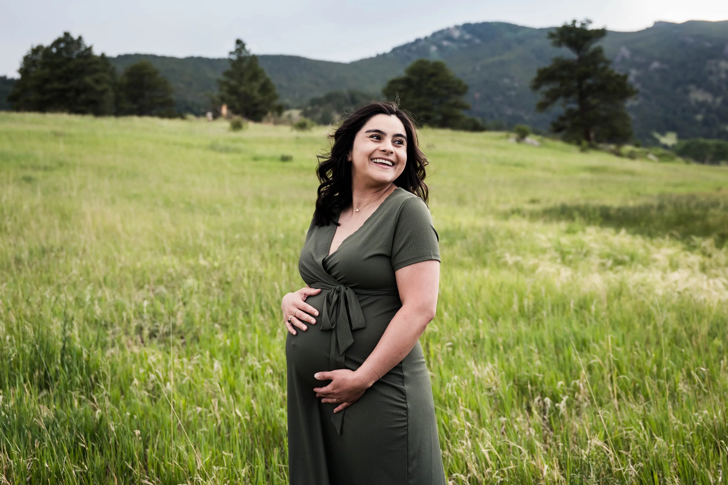 Pregnant woman standing outdoors in a green field with mountains and trees in the background, smiling and holding her belly.