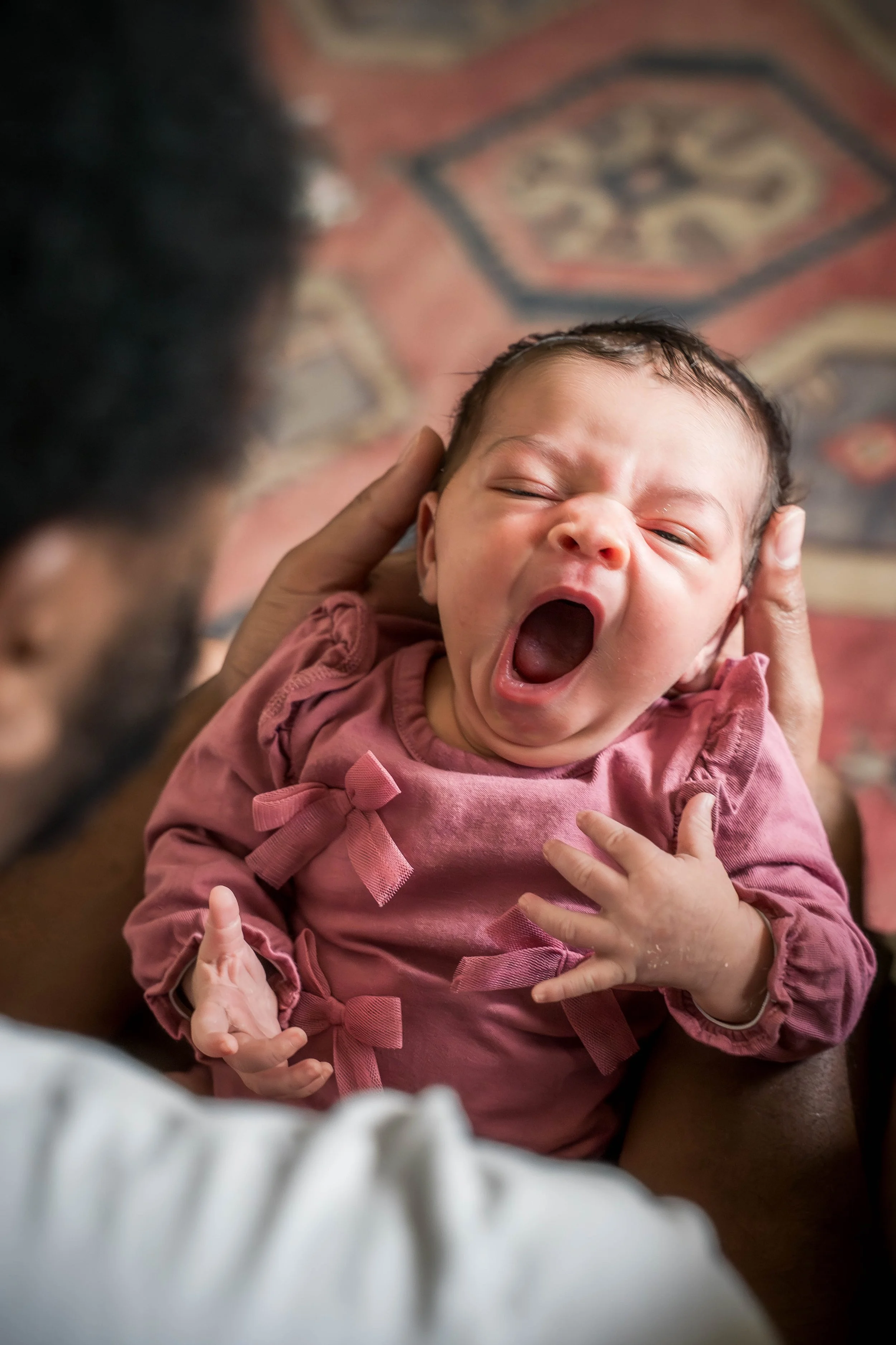 A person holding a yawning baby girl with dark hair and pink dress.