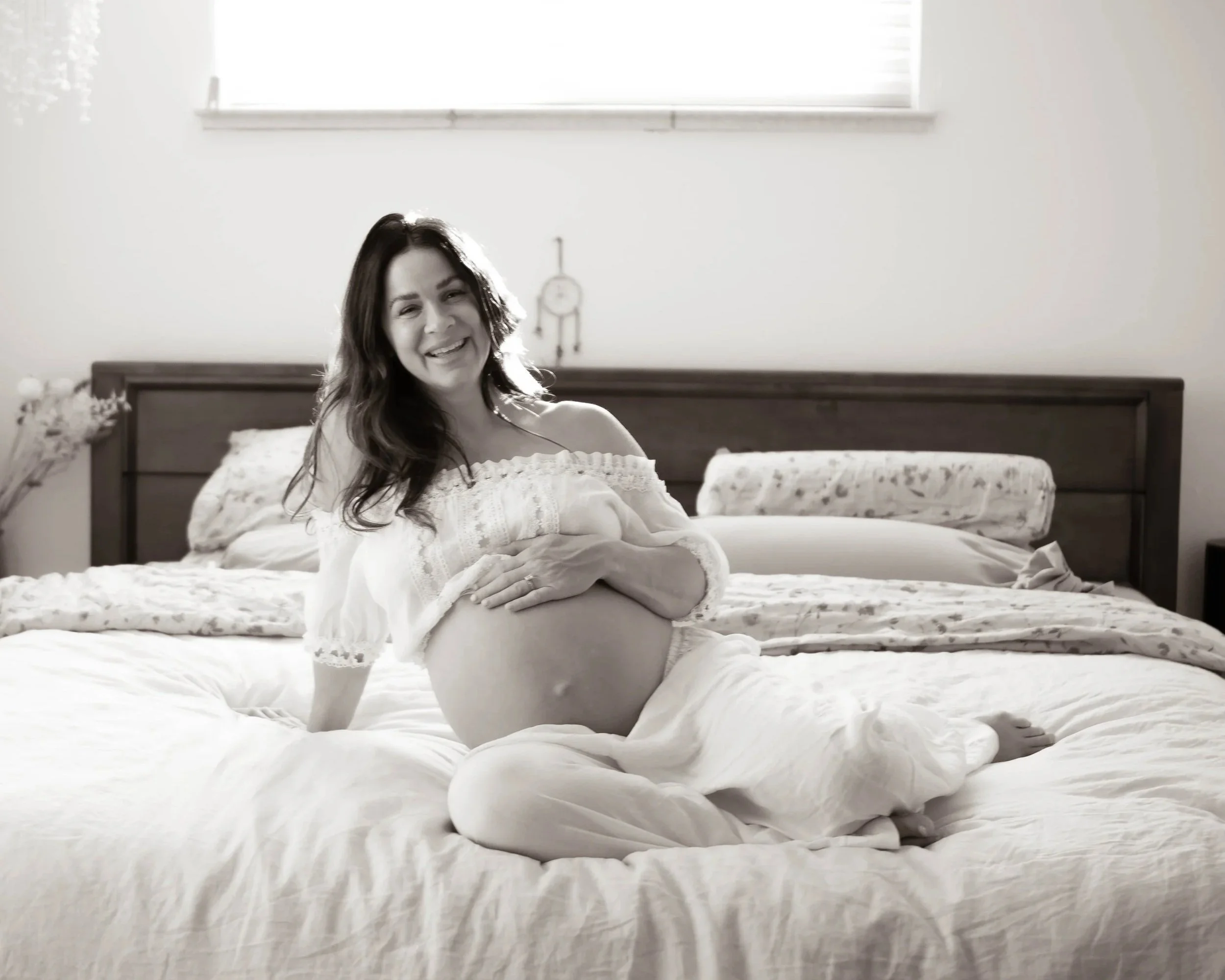 A smiling woman with dark hair in a white off-shoulder top sitting on a bed with white bedding in a bright room with a window and a dreamcatcher on the wall.