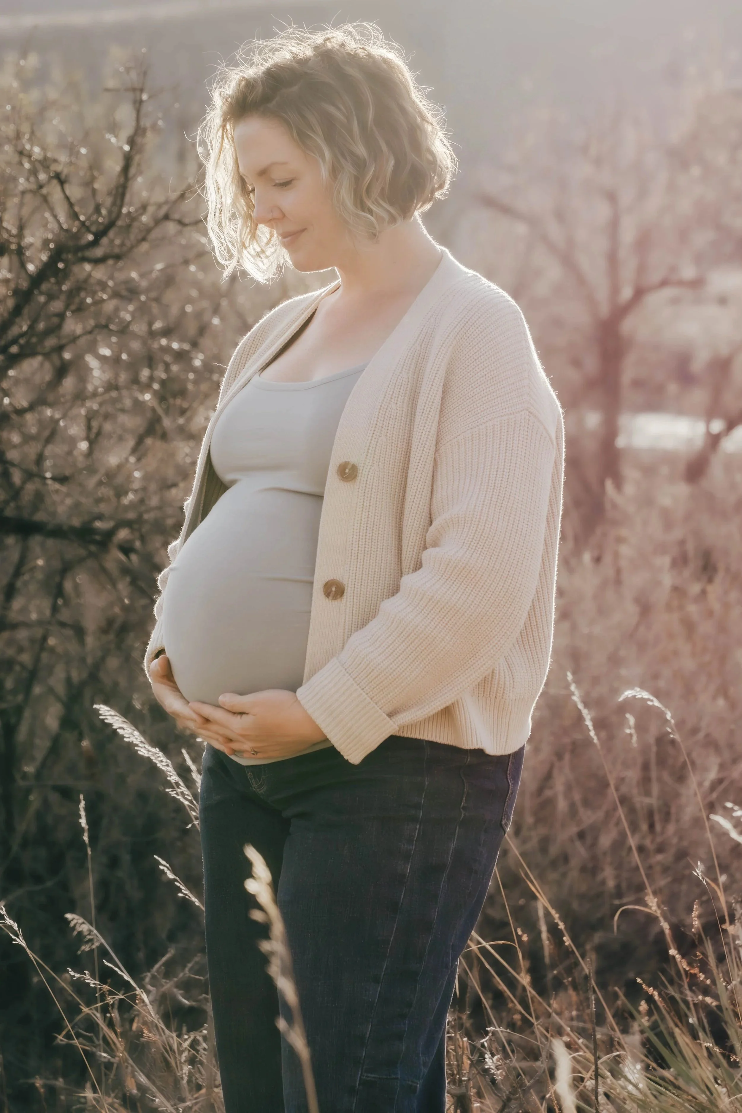 A pregnant woman with curly blonde hair stands outdoors in a field with tall grass and leafless trees in the background. She is wearing a light-colored cardigan and a white top, gently holding her belly, and smiling softly.