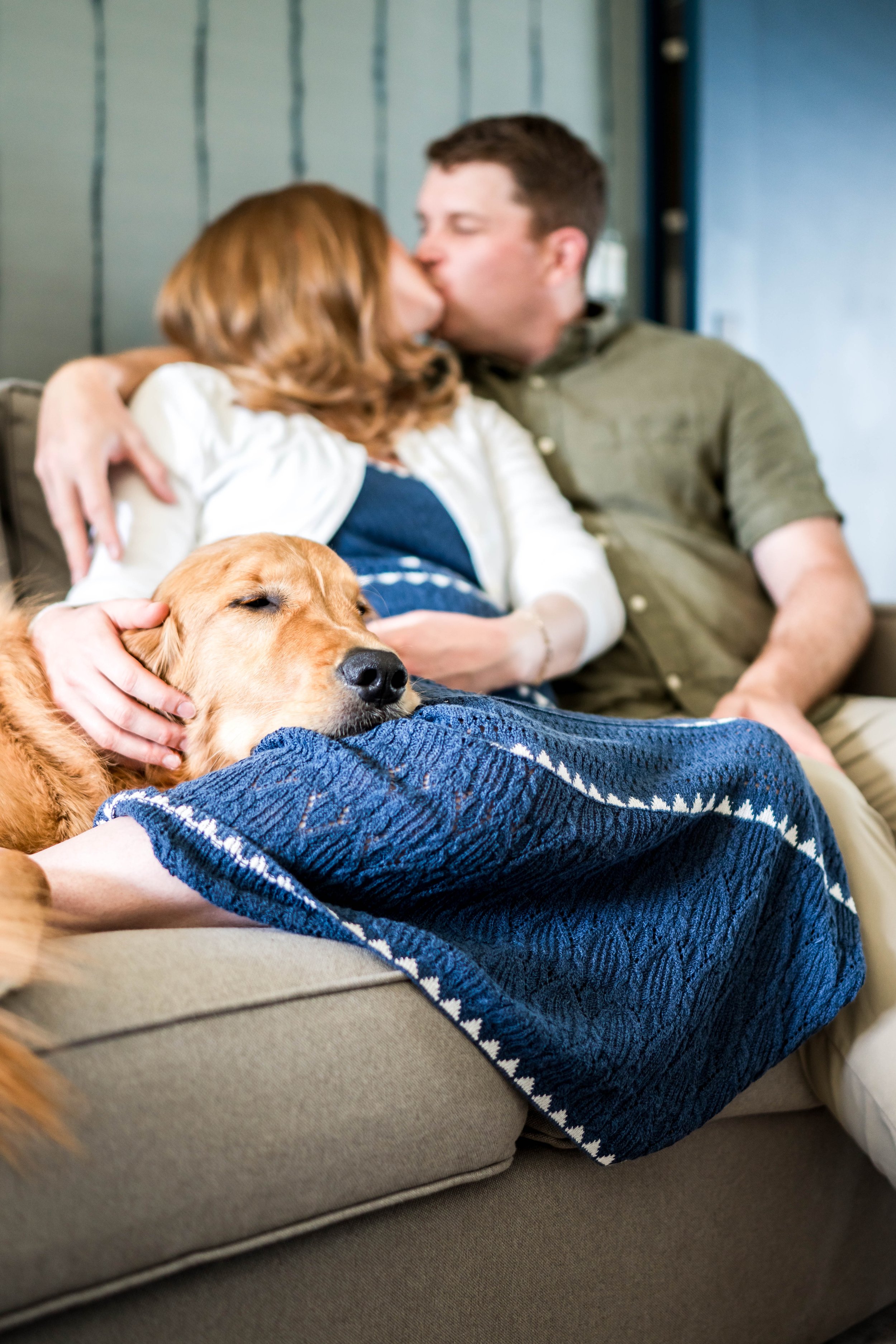 A couple sharing a kiss on a beige couch with a dog resting on their lap, covered with a blue patterned blanket. The woman has reddish-brown hair, and the man has short dark hair, wearing an olive green shirt.