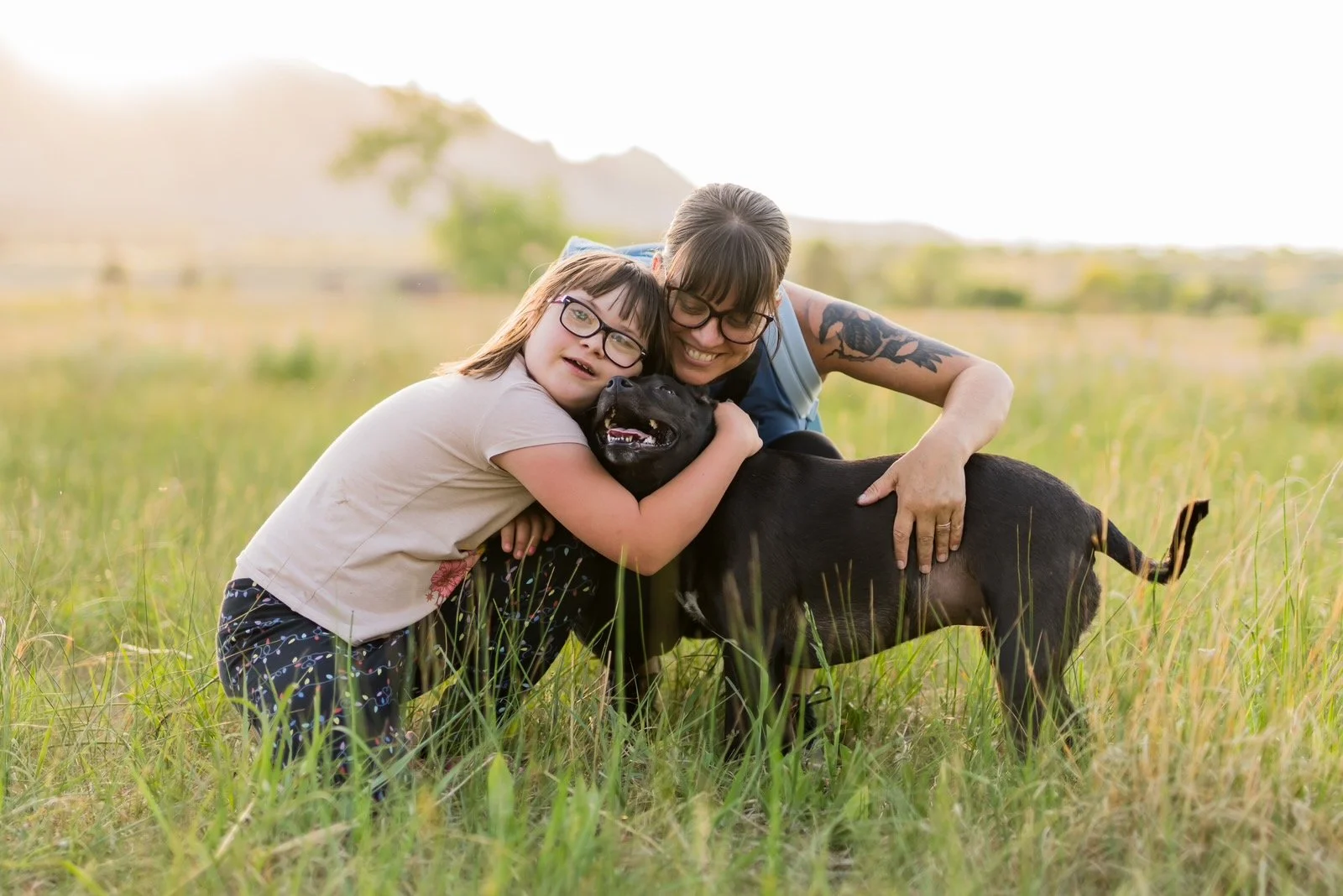 A woman and a young girl with down syndrome hugging a black dog in a grassy field in Colorado, with green trees and hills in the background on a sunny day.