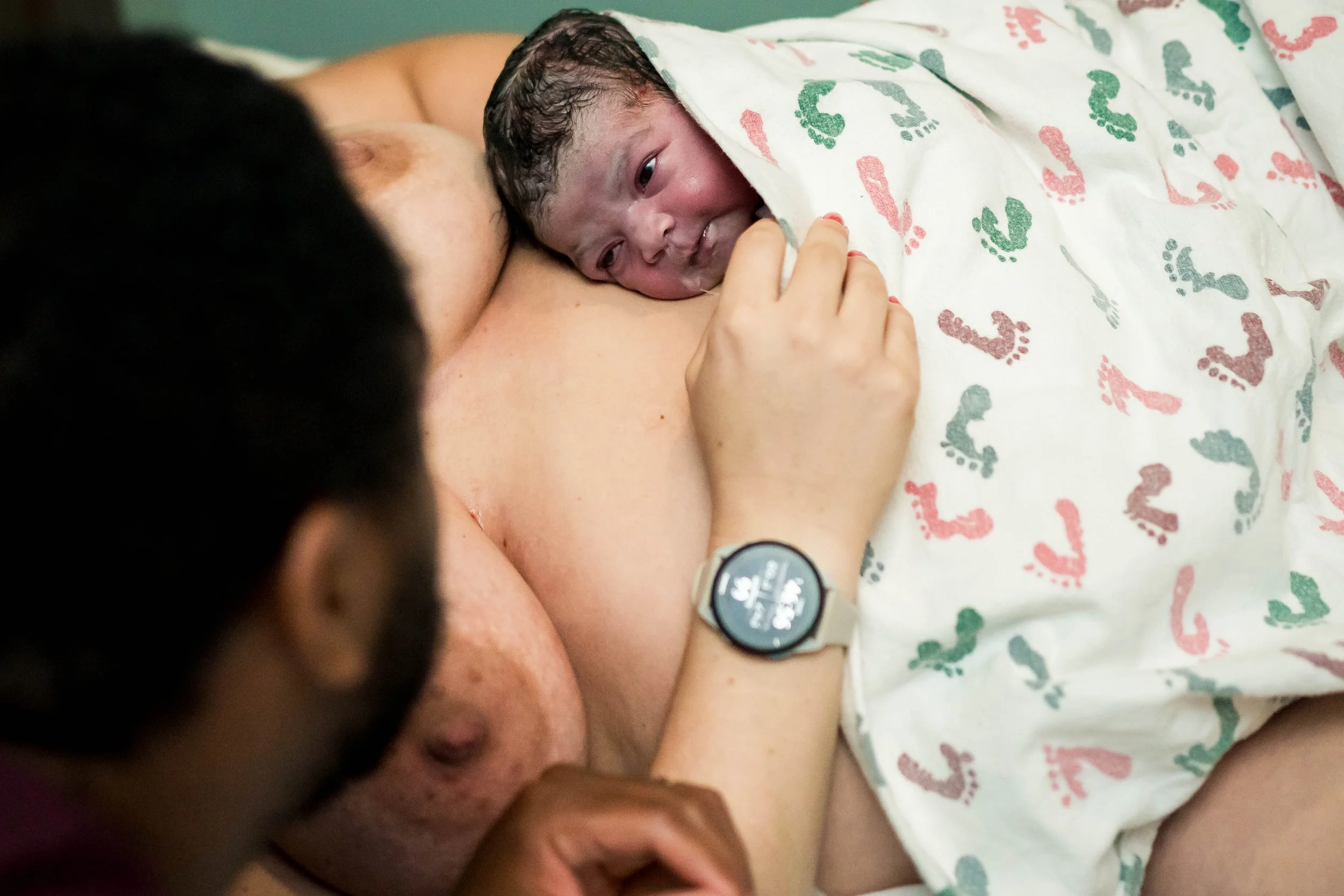 A newborn baby is resting on a person's chest, partially covered by a blanket with colorful footprints, while the person wears a watch.