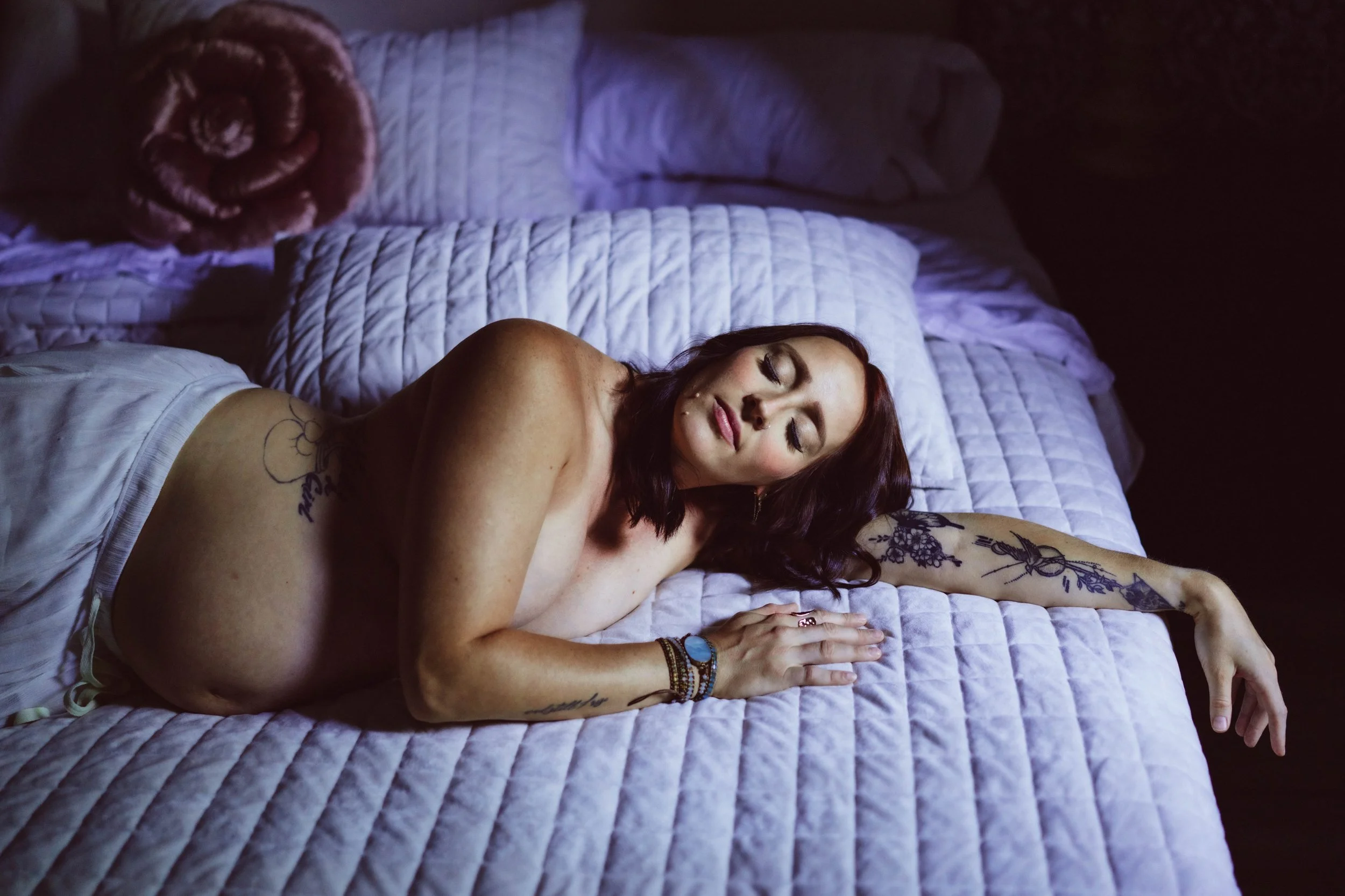 A pregnant woman with red hair and tattoos on her arm and shoulder is lying on a bed with white quilted bedding. She has closed eyes and appears to be sleeping or resting. The room is dimly lit.