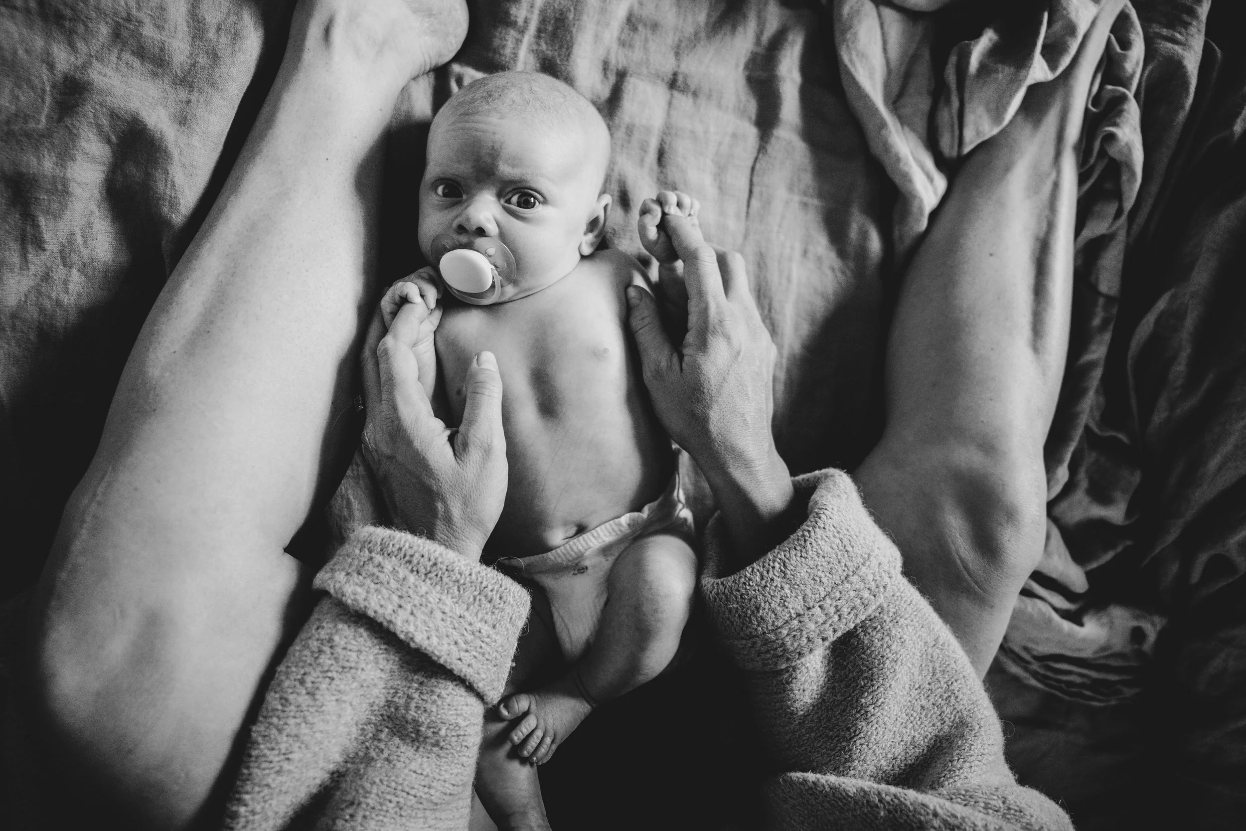 Black and white photograph of a newborn baby with a pacifier lying on a bed, surrounded by his mother's hands and arms, with some hands gently holding the baby's chest and others supporting the baby from above.
