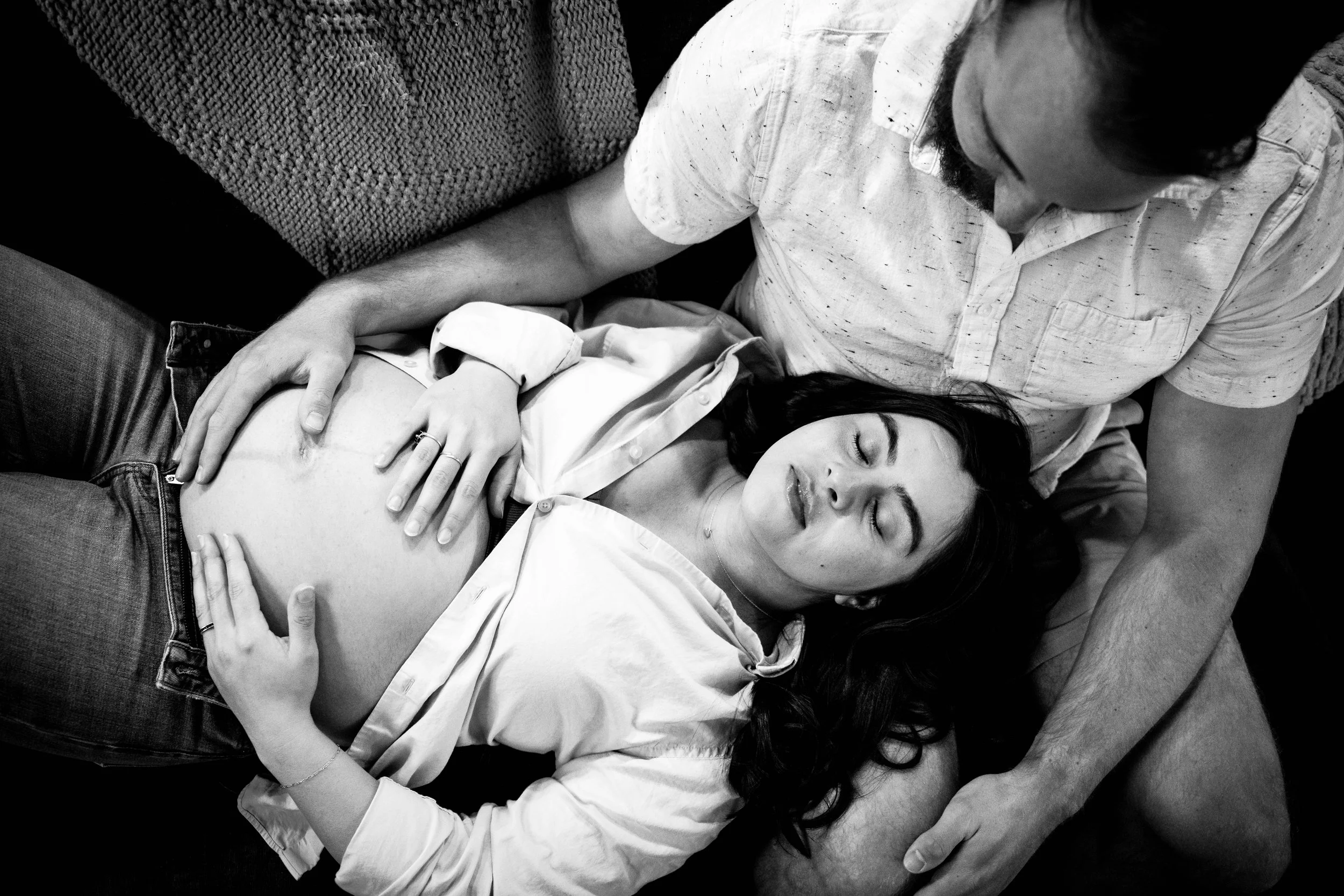 Black and white photo of a man and a pregnant woman lying on a couch, with the man holding the woman in his arms and enjoying the pregnancy.