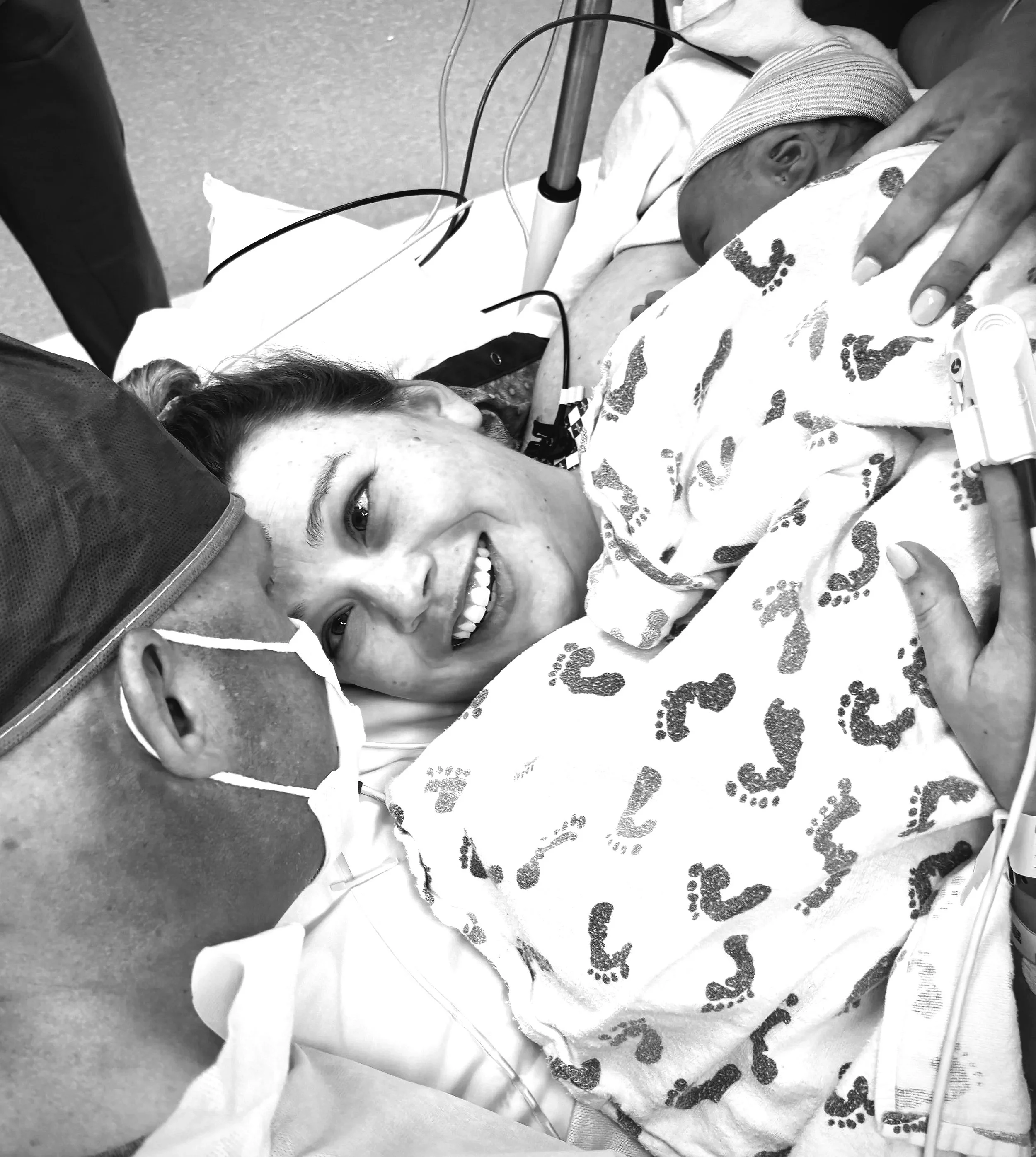 A woman lying in a hospital bed, smiling and holding a newborn baby wrapped in a hospital blanket, with medical tubes attached to the baby.