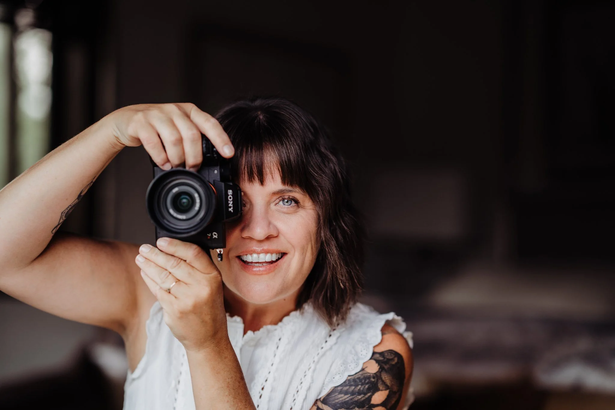 A woman with dark hair and tattoo on her arm smiling and taking a photo with a camera.