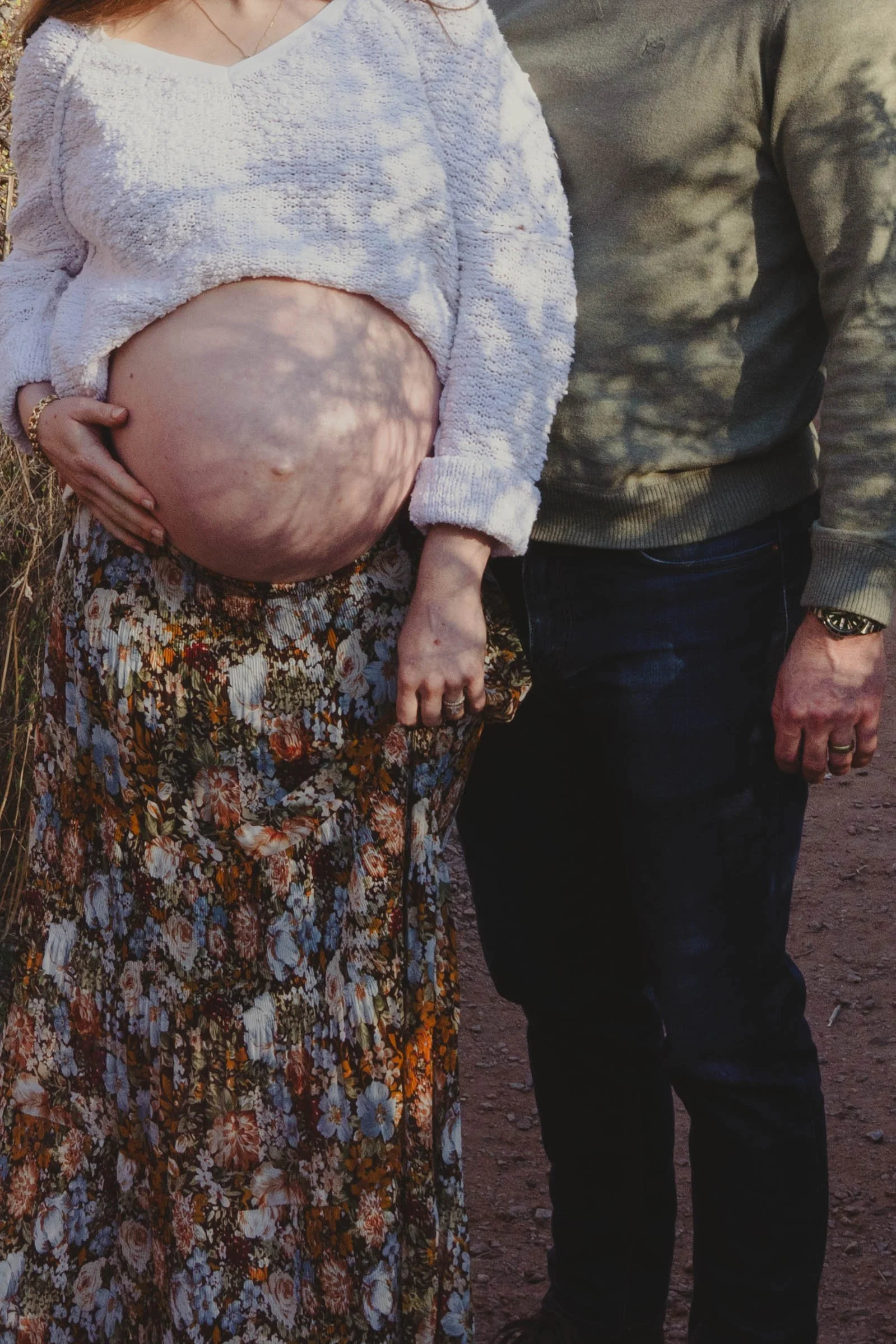 A pregnant woman wearing a floral skirt and white sweater, with her hand on her belly, standing next to a man in a gray sweater, with their bodies partially visible in an outdoor setting.
