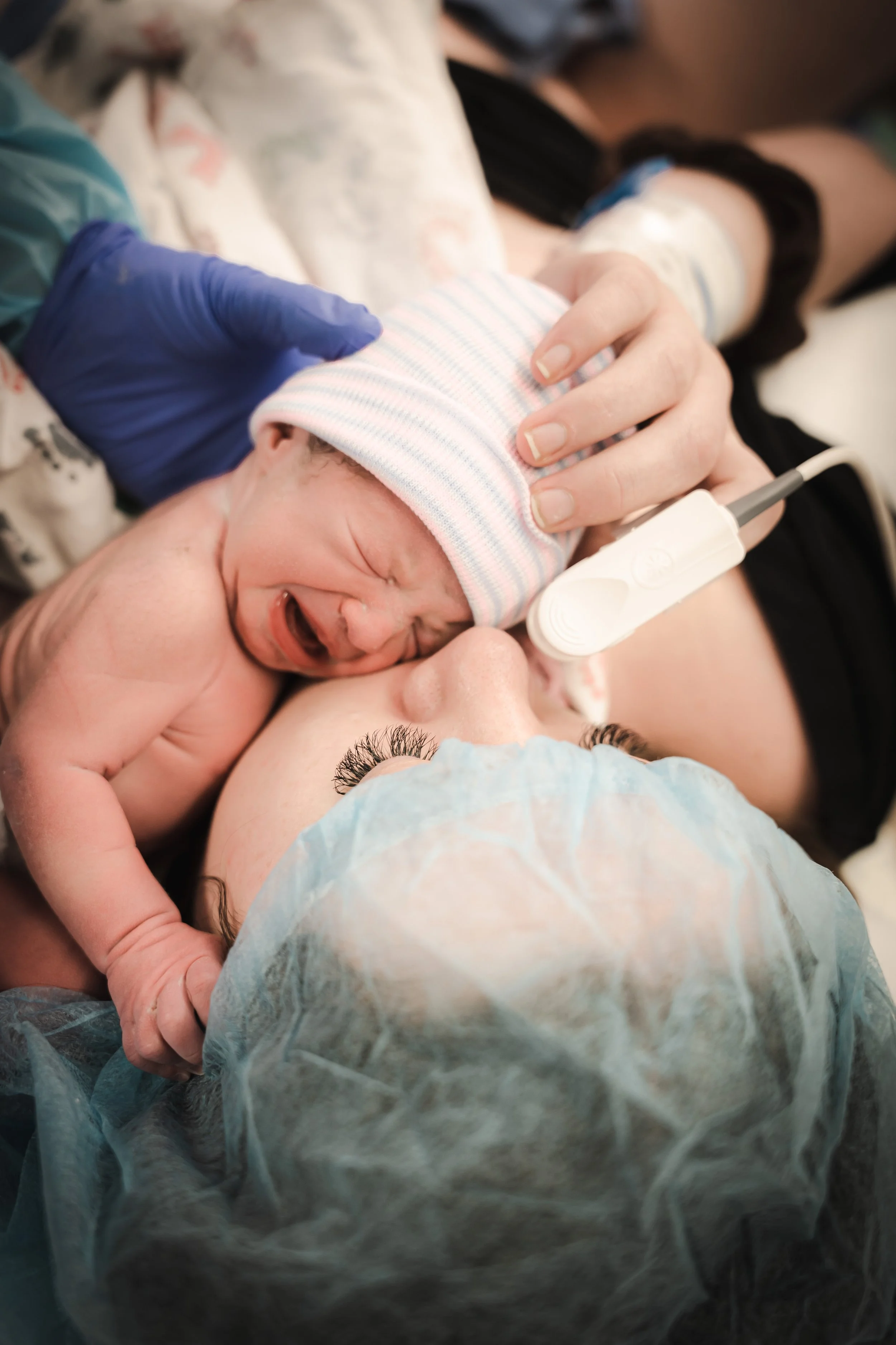 A newborn baby lying on a hospital bed, crying while being gently held by a person. The baby is wearing a pink and blue striped hospital hat, and there is medical equipment connected to the baby, including a sensor on the foot. The setting appears to