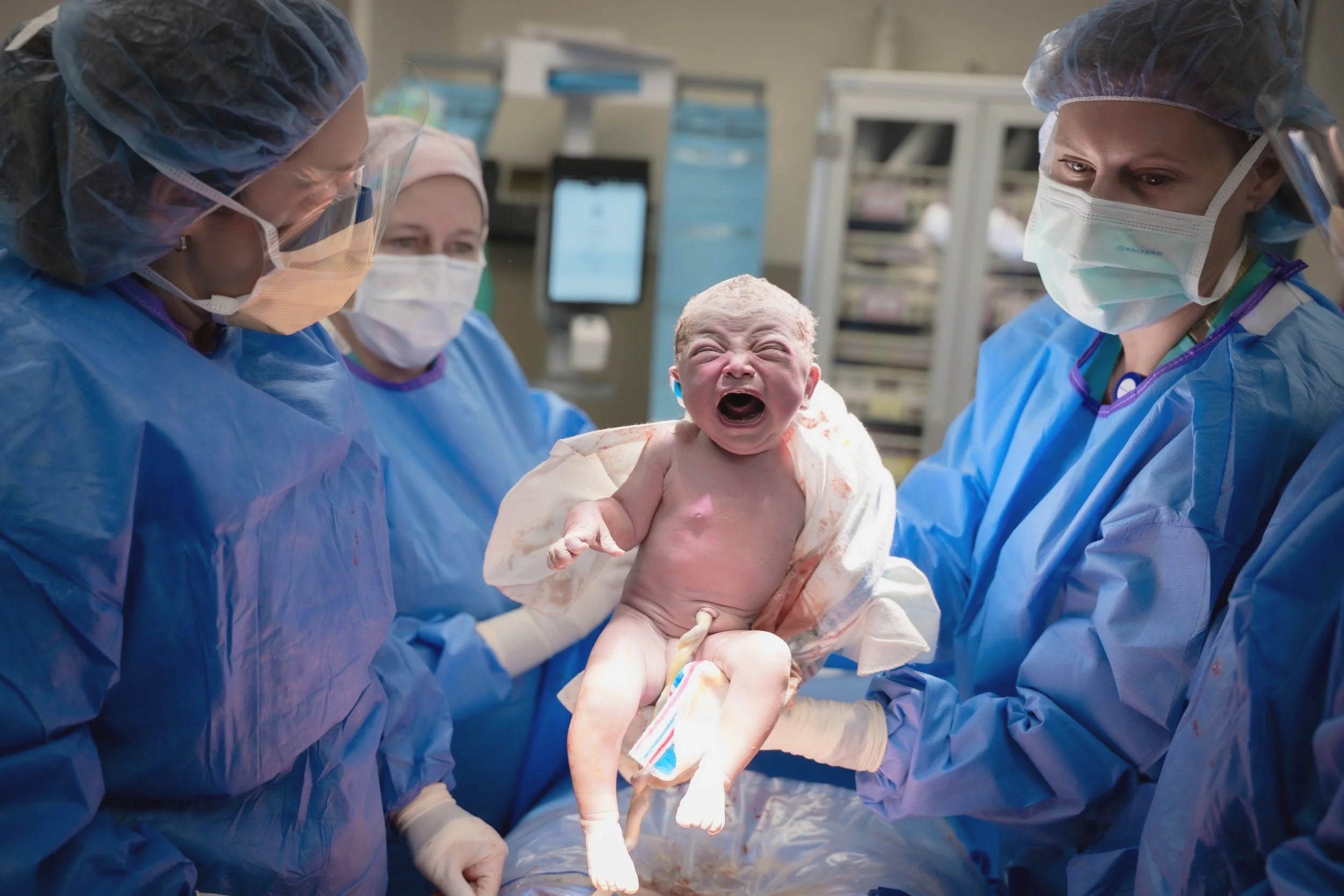 Medical professionals in blue scrubs, masks, and hair covers assisting in delivery of a baby in a hospital setting.
