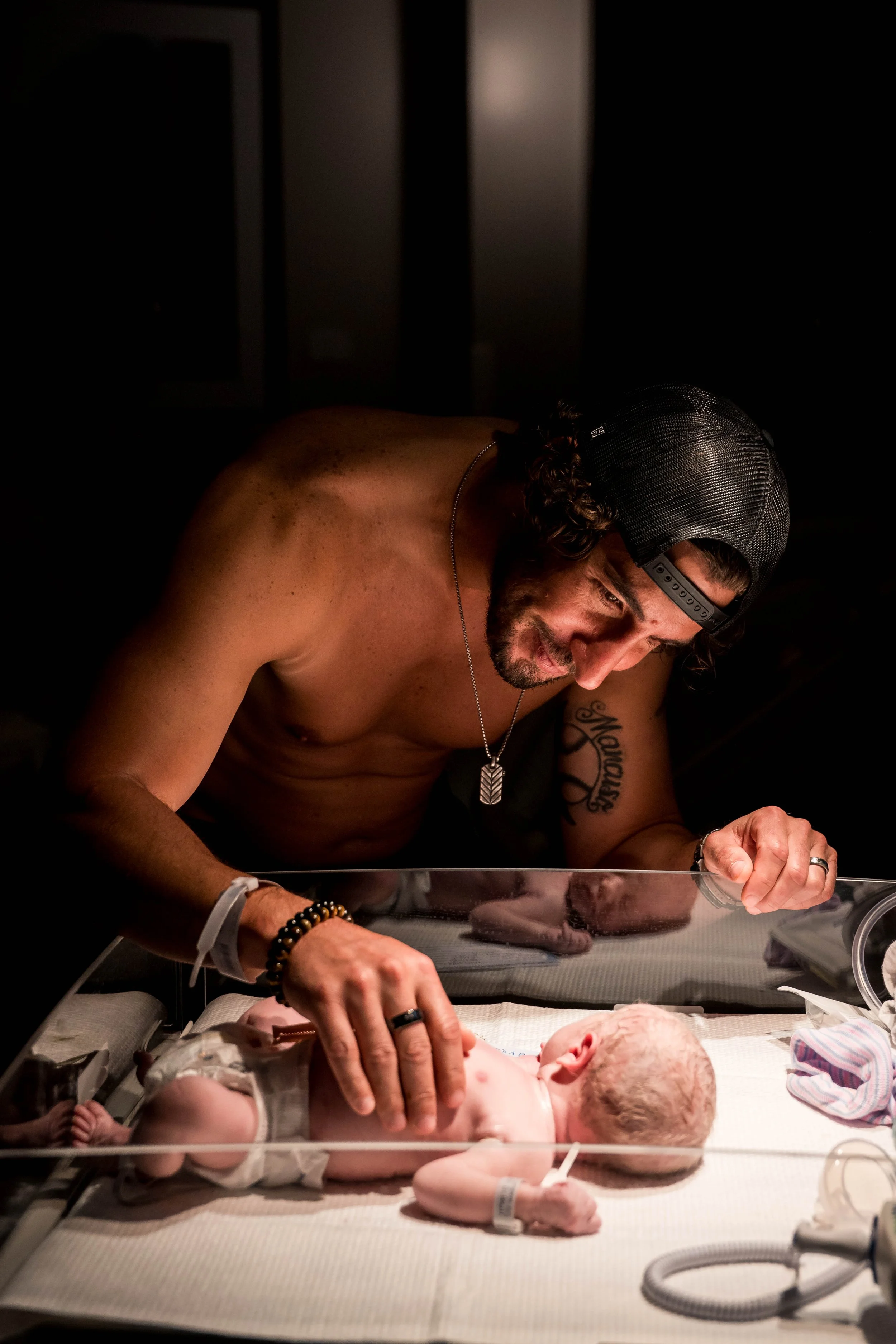 A man with a beard, wearing a black cap and jewelry, leans over an infant on a hospital bassinet, holding the baby gently. The baby appears to be newborn, lying on its back in a hospital setting with medical equipment nearby.