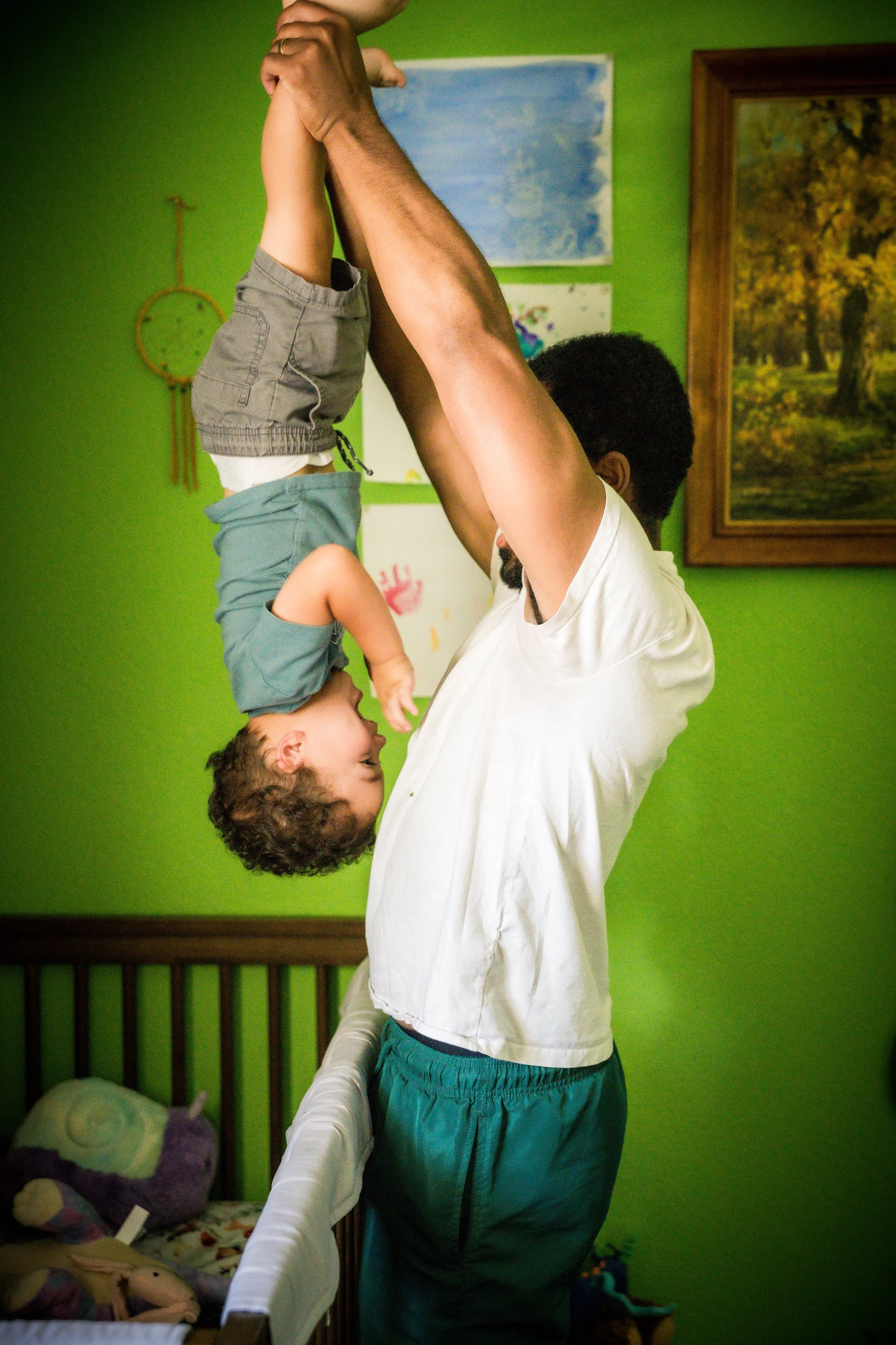 A man lifting a young child upside down in a brightly colored green room with artwork and a bed with stuffed animals.