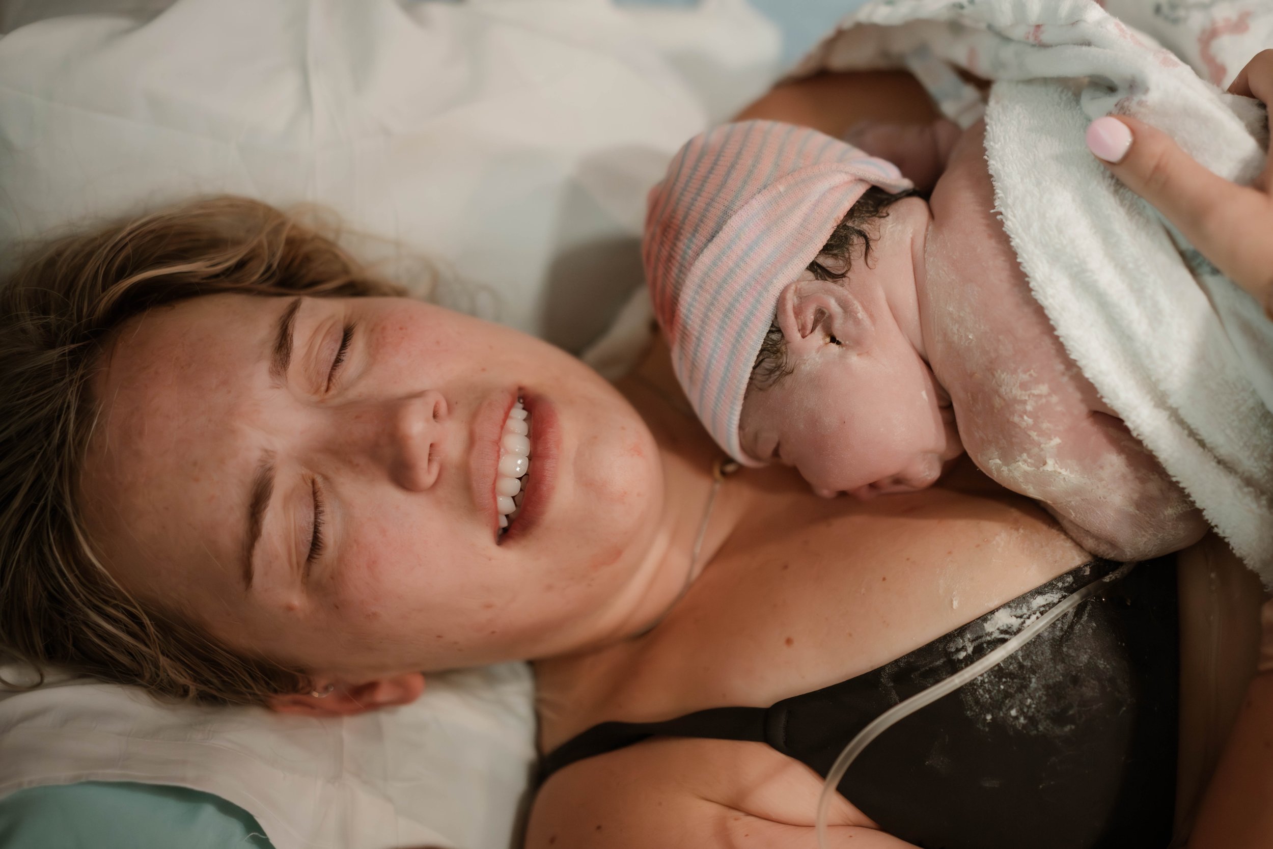 A woman with curly blond hair is lying in a hospital bed, smiling, holding a newborn baby wrapped in a blanket. The baby was just born and the mother is relieved and grateful.