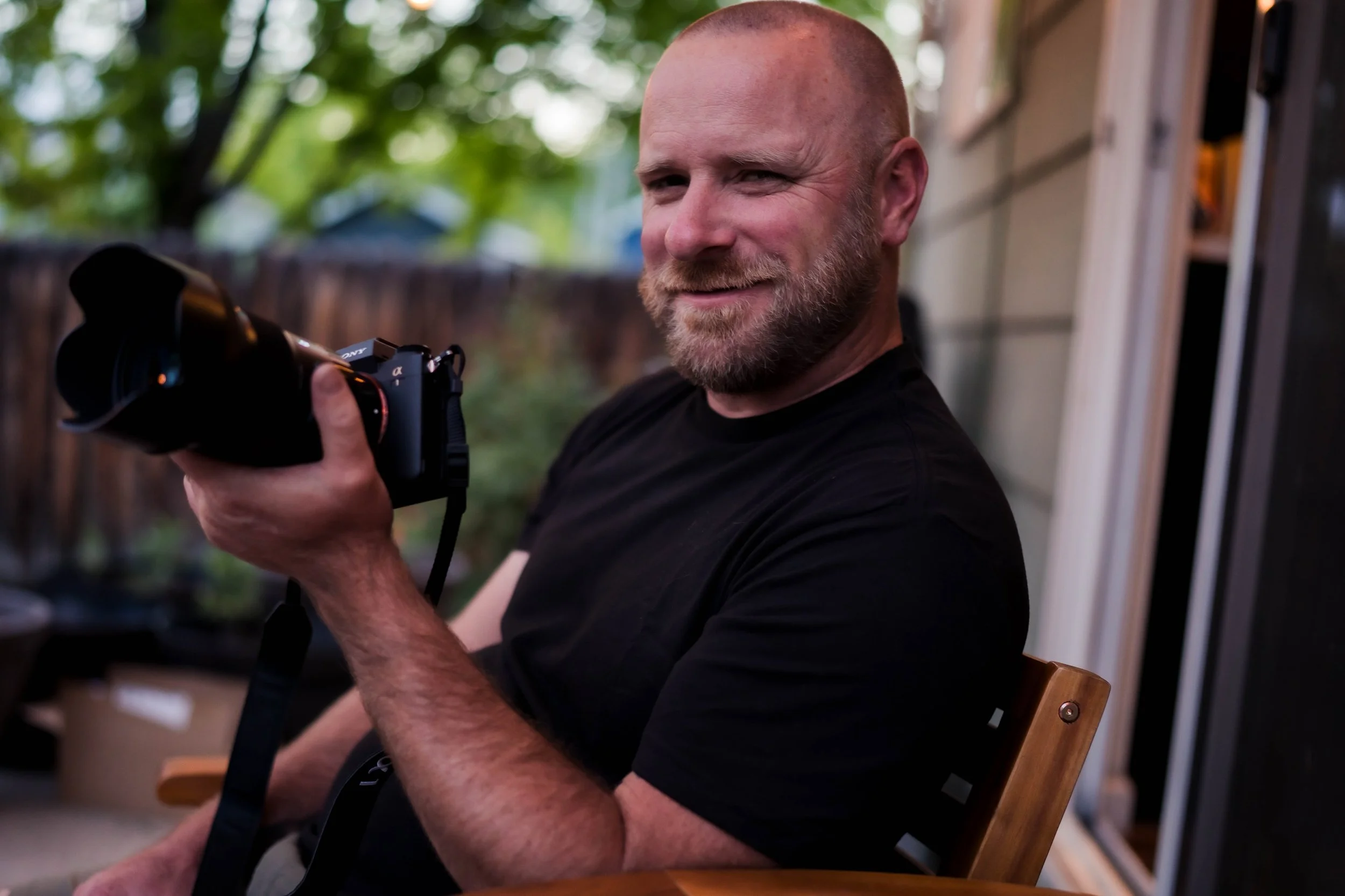 A smiling man with a beard holding a camera, sitting on a wooden chair outdoors with trees and a house in the background.