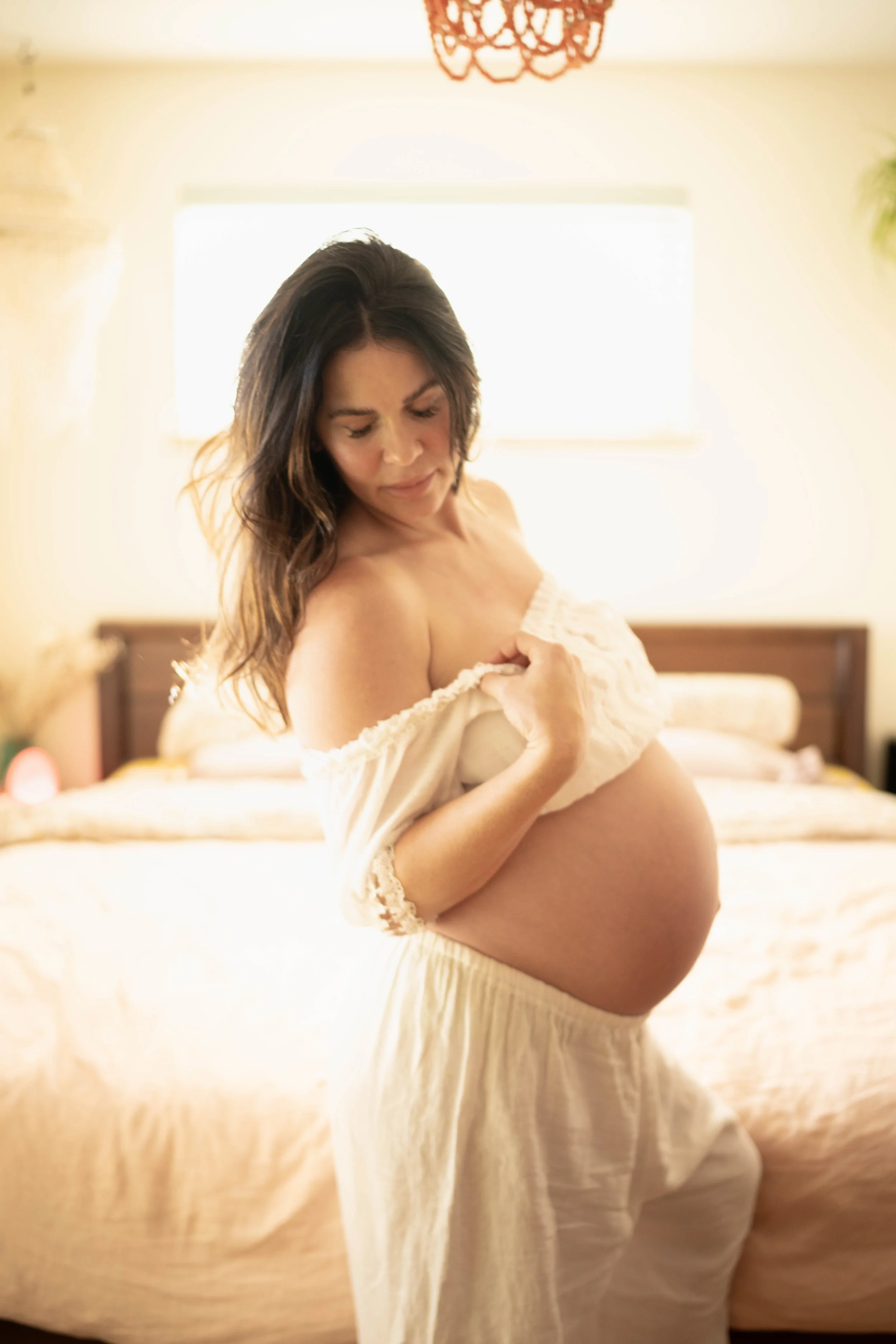 A pregnant woman with long dark hair looking down, holding her white off-shoulder top, in a softly lit bedroom.
