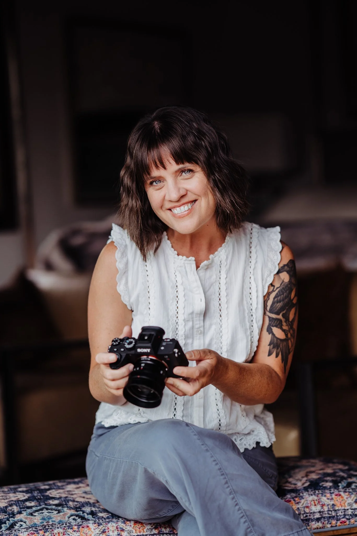 A smiling woman with dark brown, wavy hair and tattoos on her left arm, sitting on a patterned rug, holding a camera in her hands.