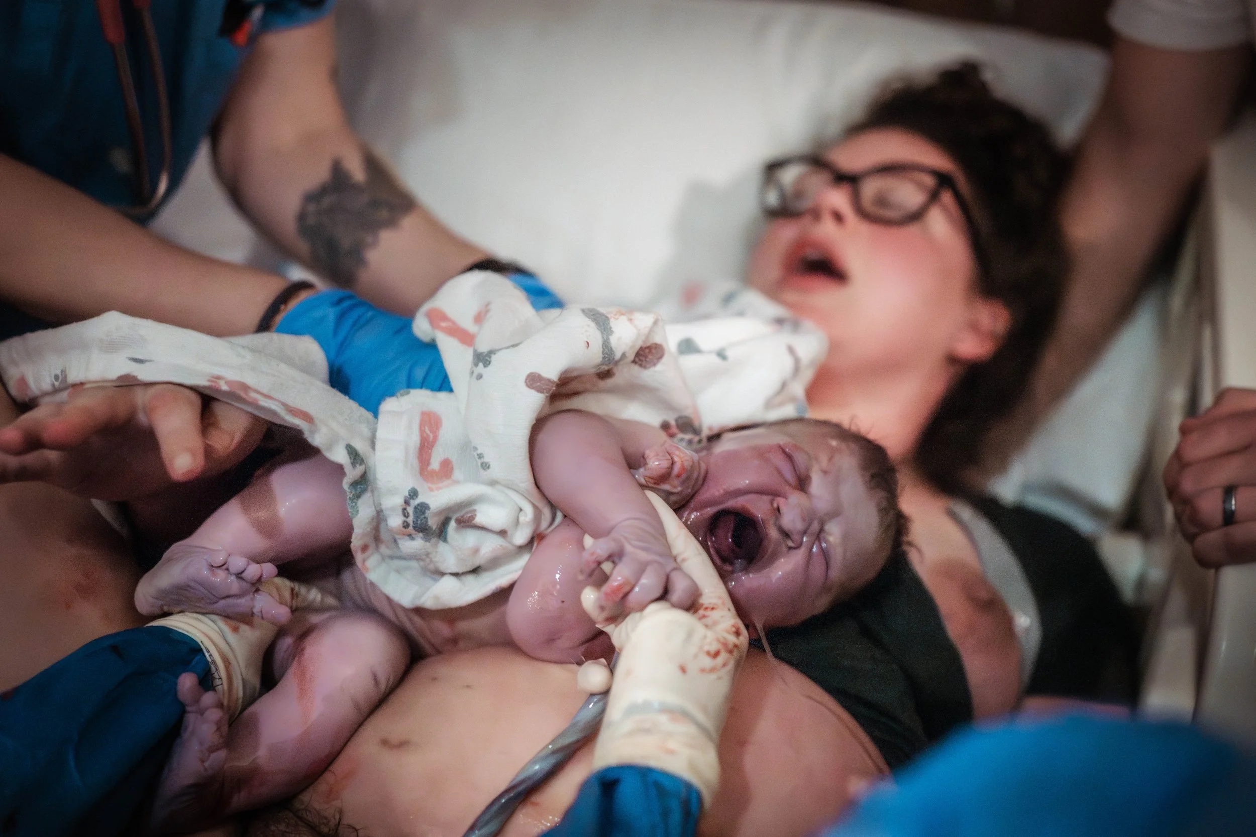 A woman giving birth to a baby in a hospital. The woman appears exhausted and is holding the newborn, which is still covered in amniotic fluid and blood.