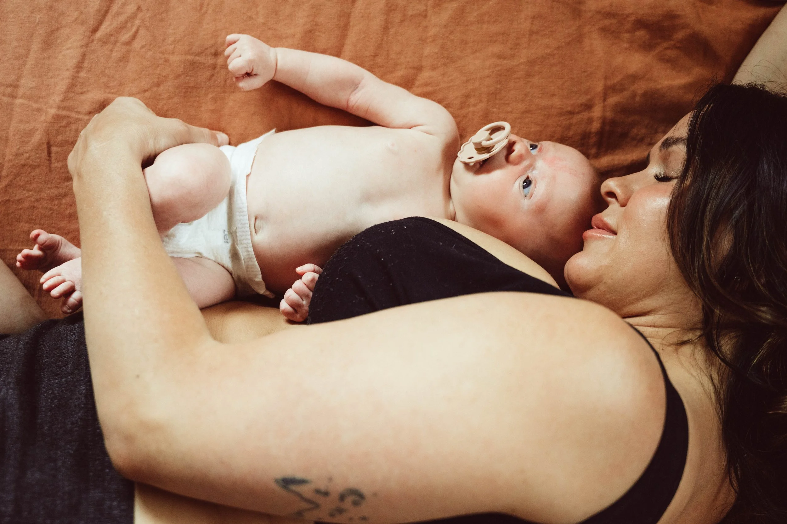 A woman lying on her side on a brown surface, looking at a baby who is lying on her chest. The baby has a pacifier in its mouth and is looking up. The woman has long dark hair and a tattoo on her arm.