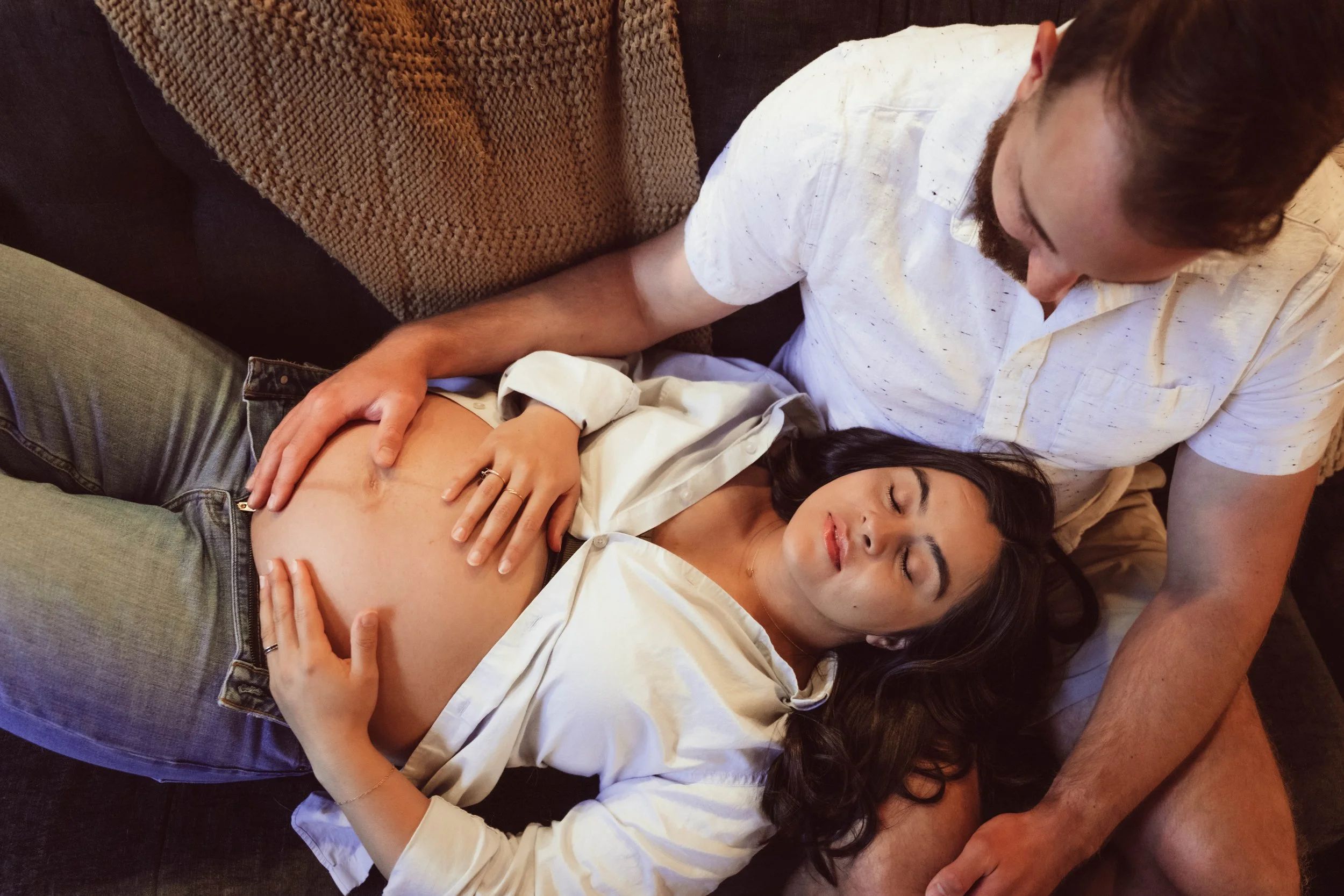 A man and woman lying on a couch, with the woman pregnant, resting and appearing to sleep.