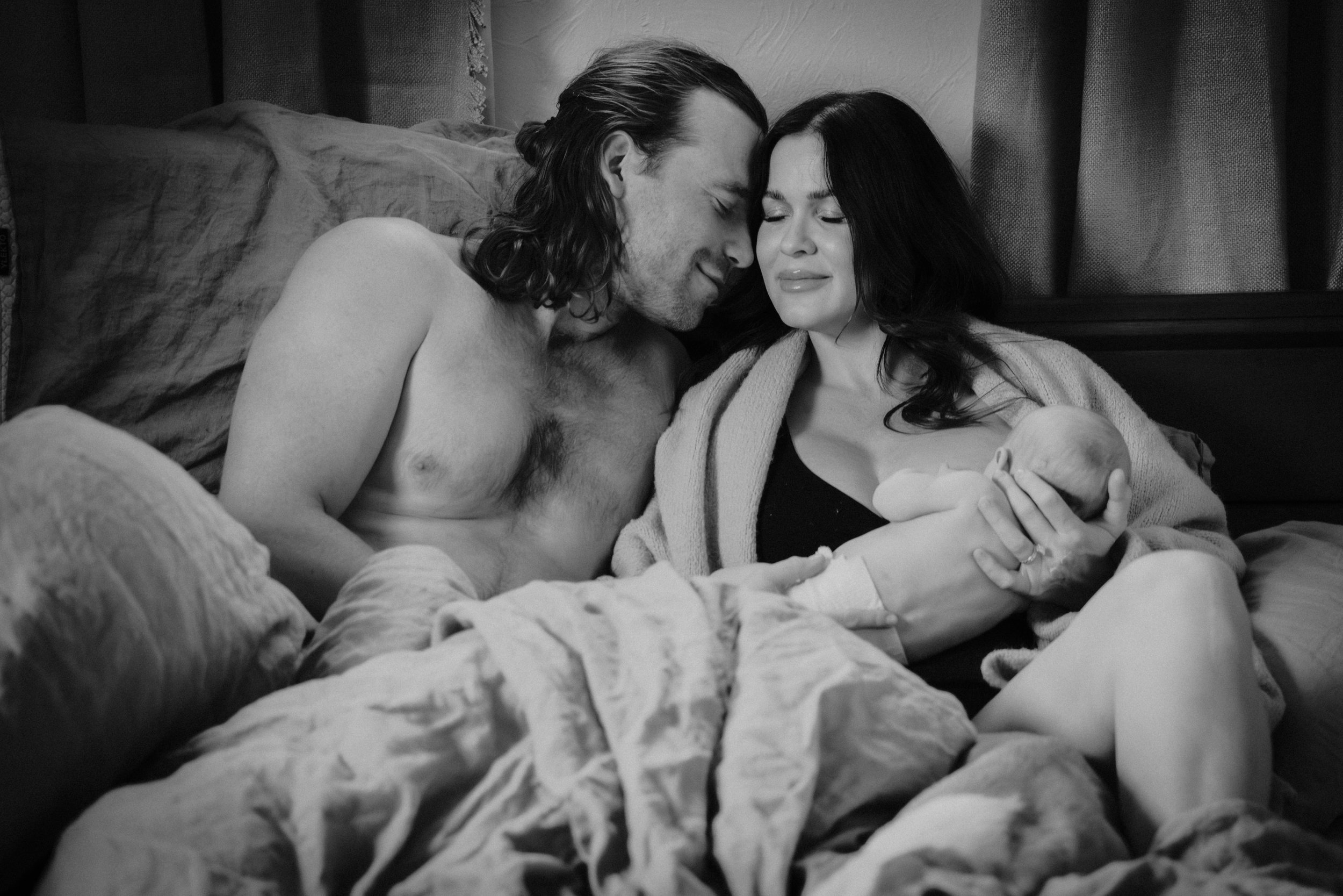 A couple with long hair and light skin sitting in bed, smiling and tenderly looking at their newborn baby.