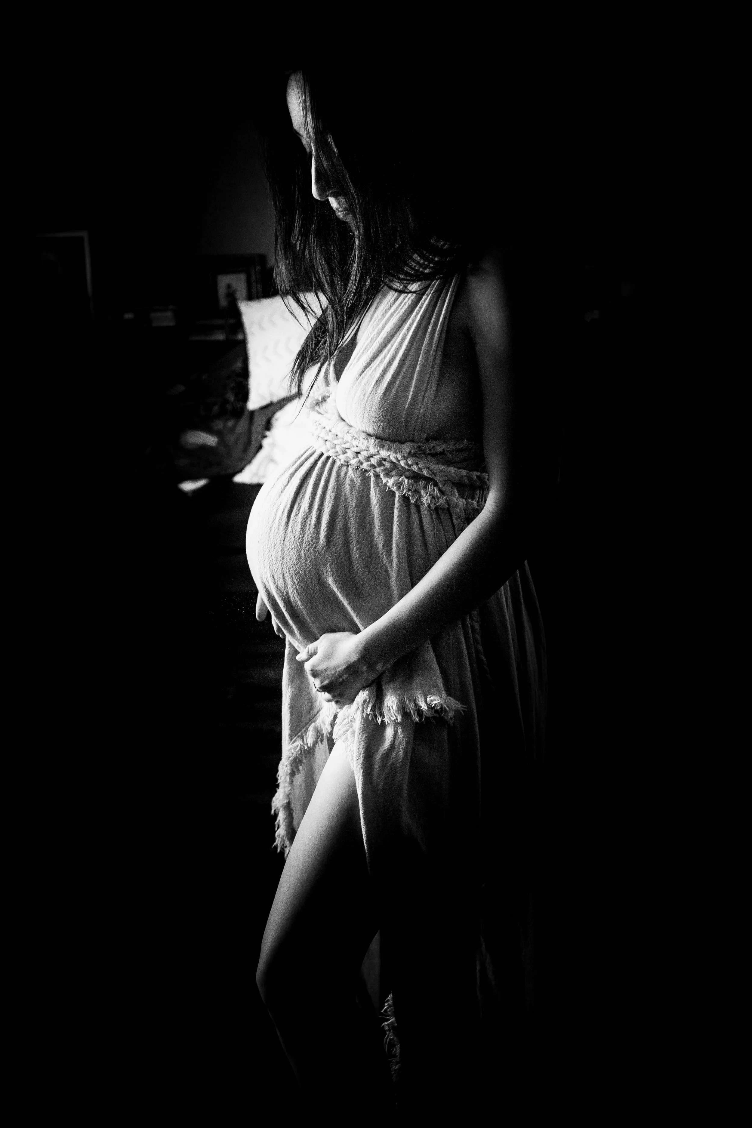 A pregnant woman in a flowing dress standing in a dark room, illuminated softly, with her hand gently resting on her belly.
