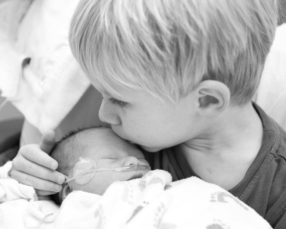 A young boy gently kissing an infant who is lying in a hospital bed with a nasal oxygen tube.