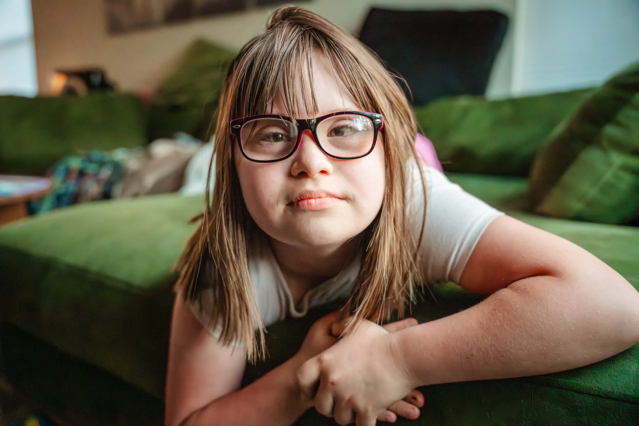 A young girl with down syndrome and glasses lying on a green couch smiling, with a blurred background including a stool and plants.