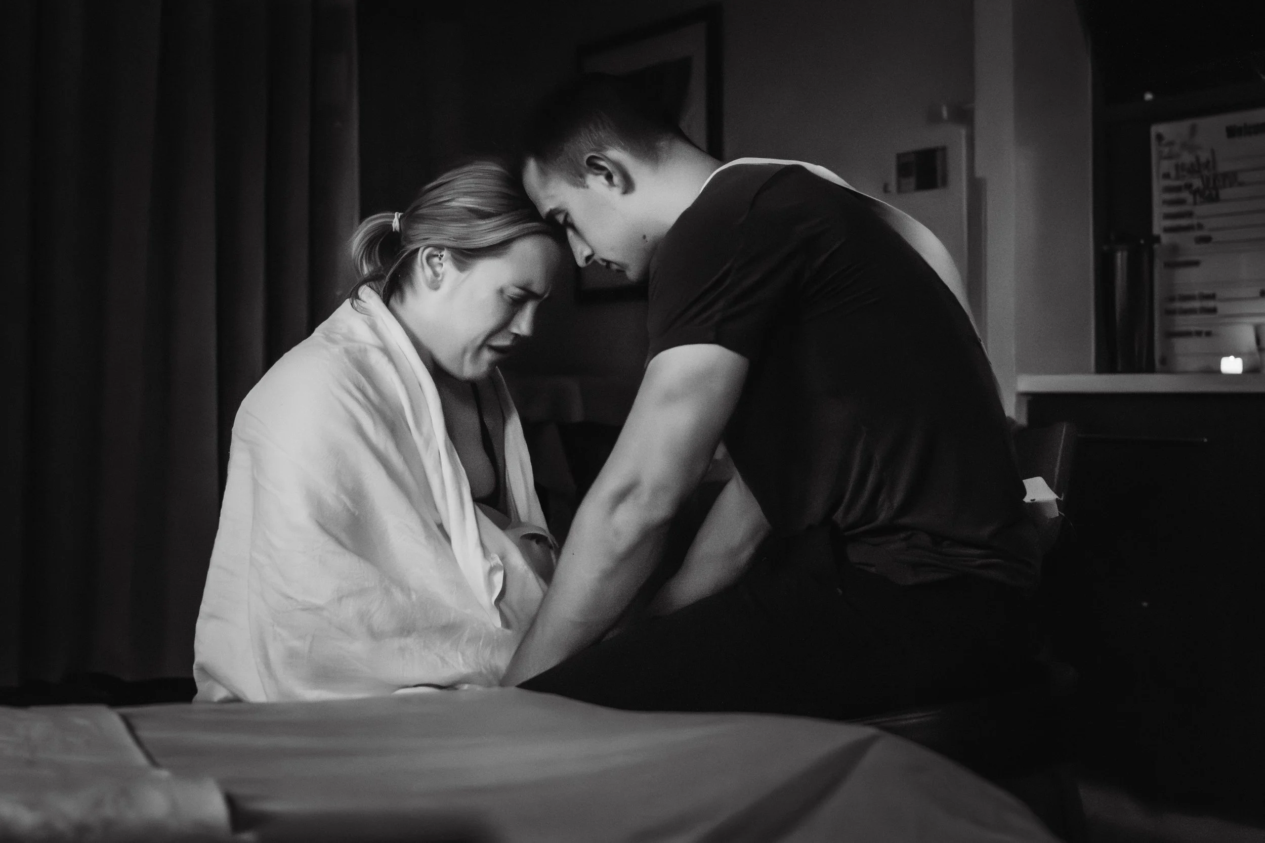 A woman with blonde hair in a ponytail, wearing a white robe, and a man with short dark hair and a dark shirt, leaning their foreheads together in a tender moment in a dimly lit room during labor right before birth.